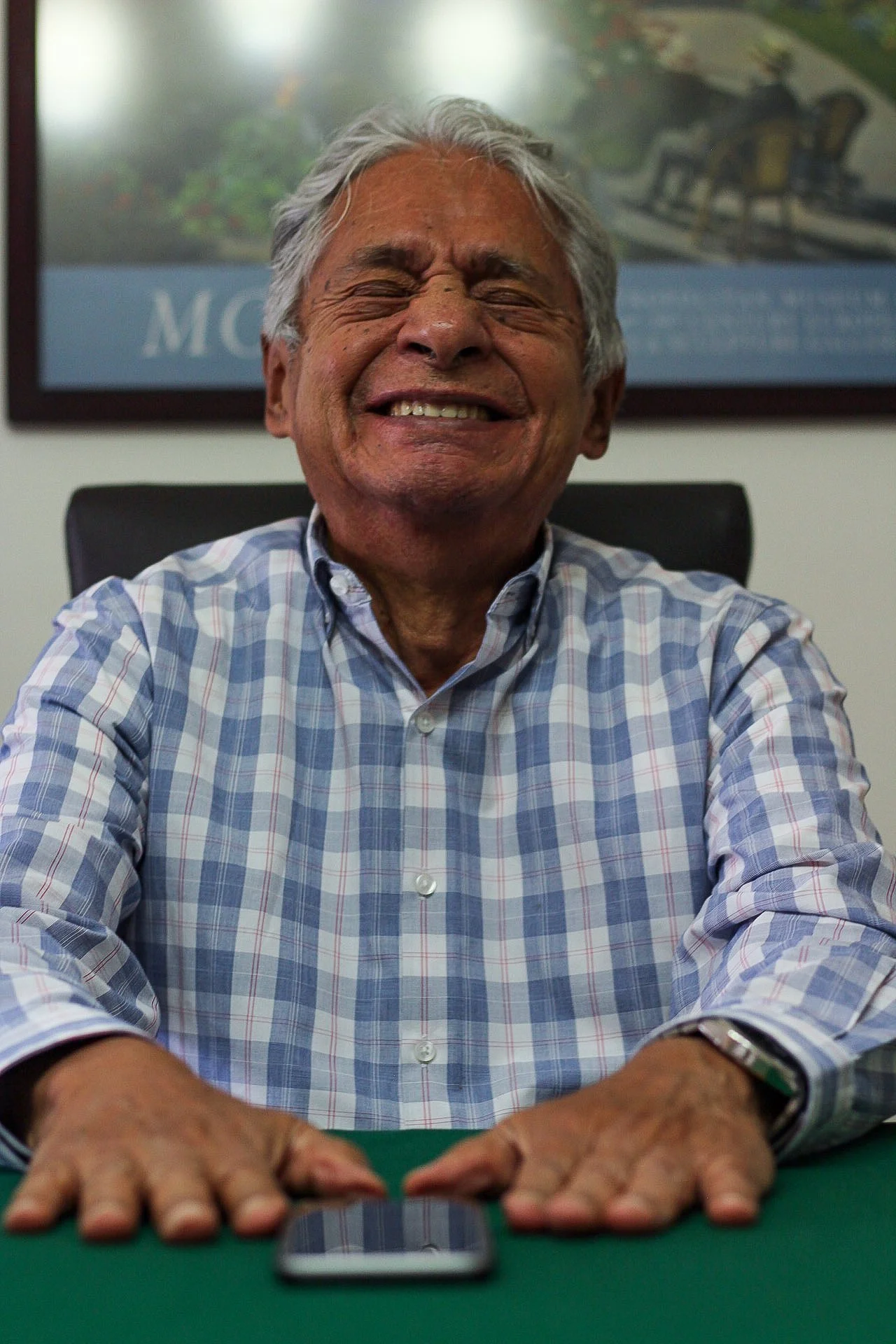 Hombre mayor con cabello gris, sonriendo con los ojos cerrados, sentado en una mesa con un teléfono móvil frente a él.