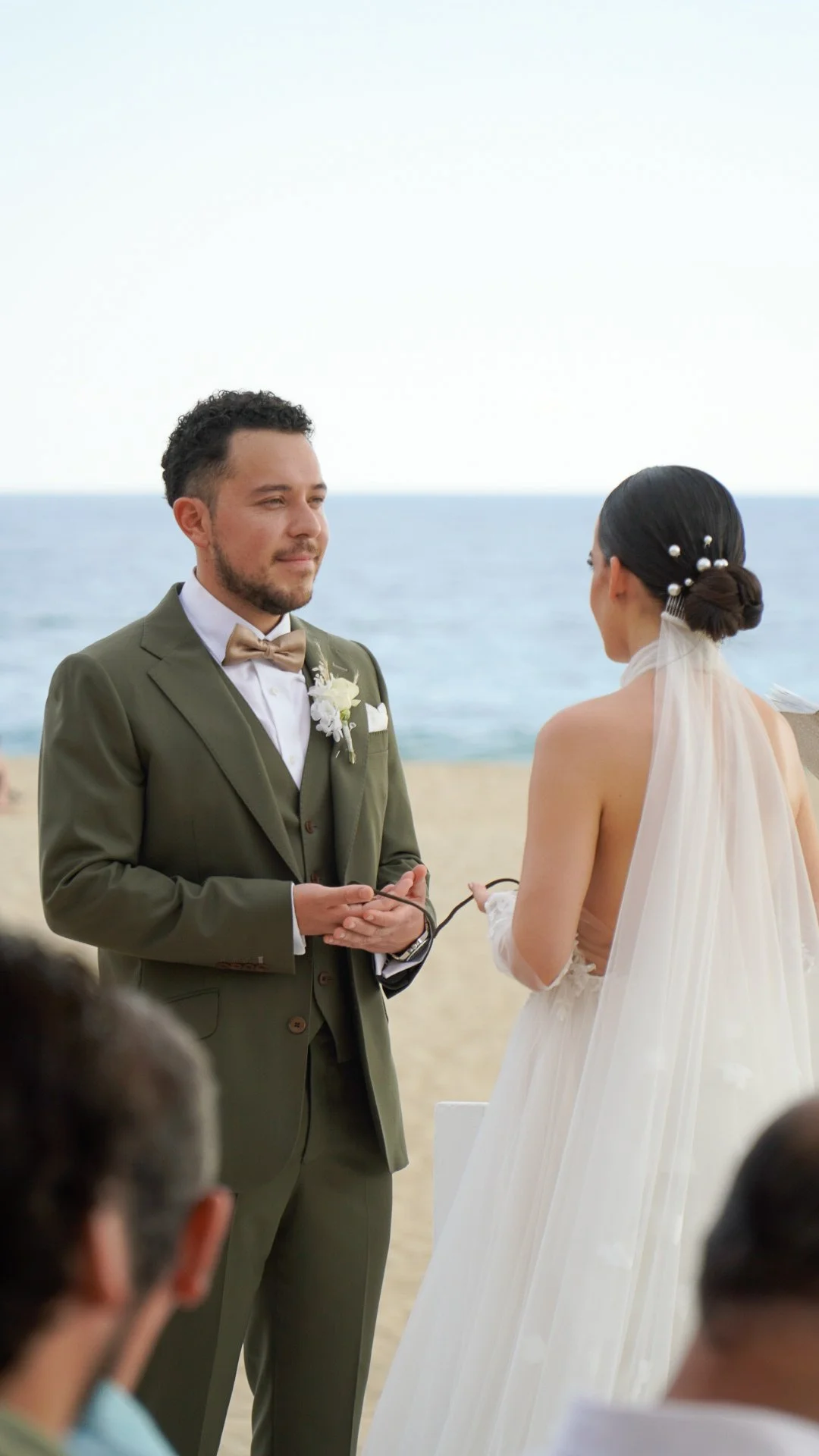 Una pareja en una ceremonia de boda en la playa, con el mar y el cielo despejado de fondo, la mujer con un vestido blanco y el hombre con traje verde, ambos de espaldas.