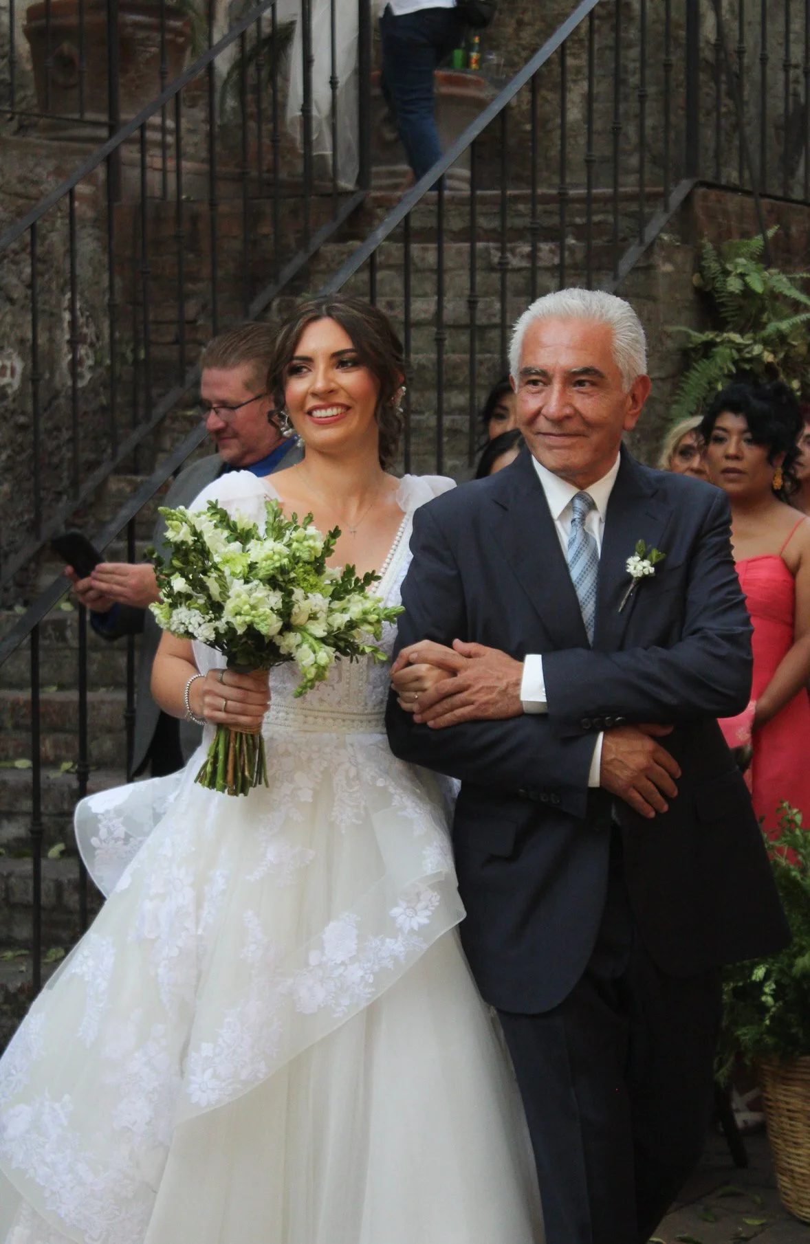 Una novia con vestido blanco y un ramo de flores, acompañada por un hombre mayor, probablemente su padre, en una ceremonia de boda en un entorno al aire libre con escalones de piedra y un grupo de invitados en el fondo.