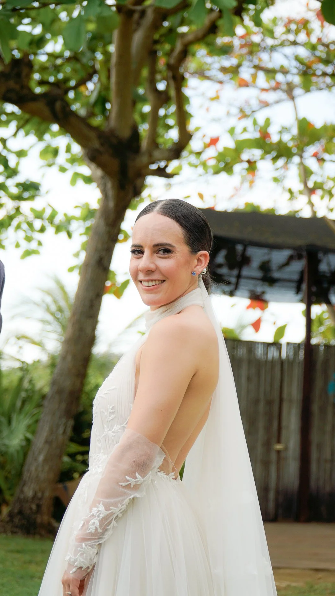 Mujer en vestido de novia sonriendo al aire libre con árboles y estructura de fondo.