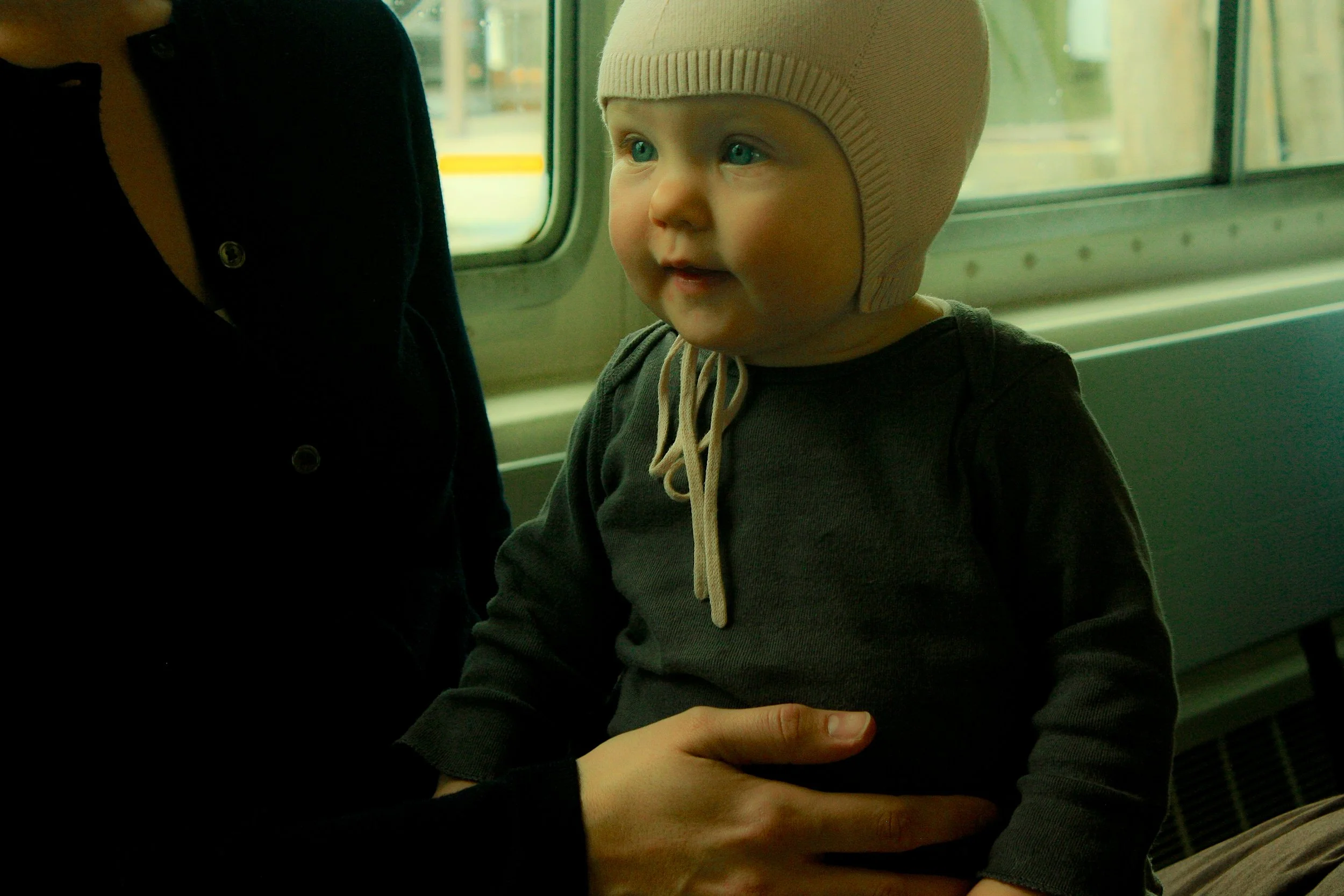 Niño con gorro beige sentado en un transporte público, sostenido por una persona.