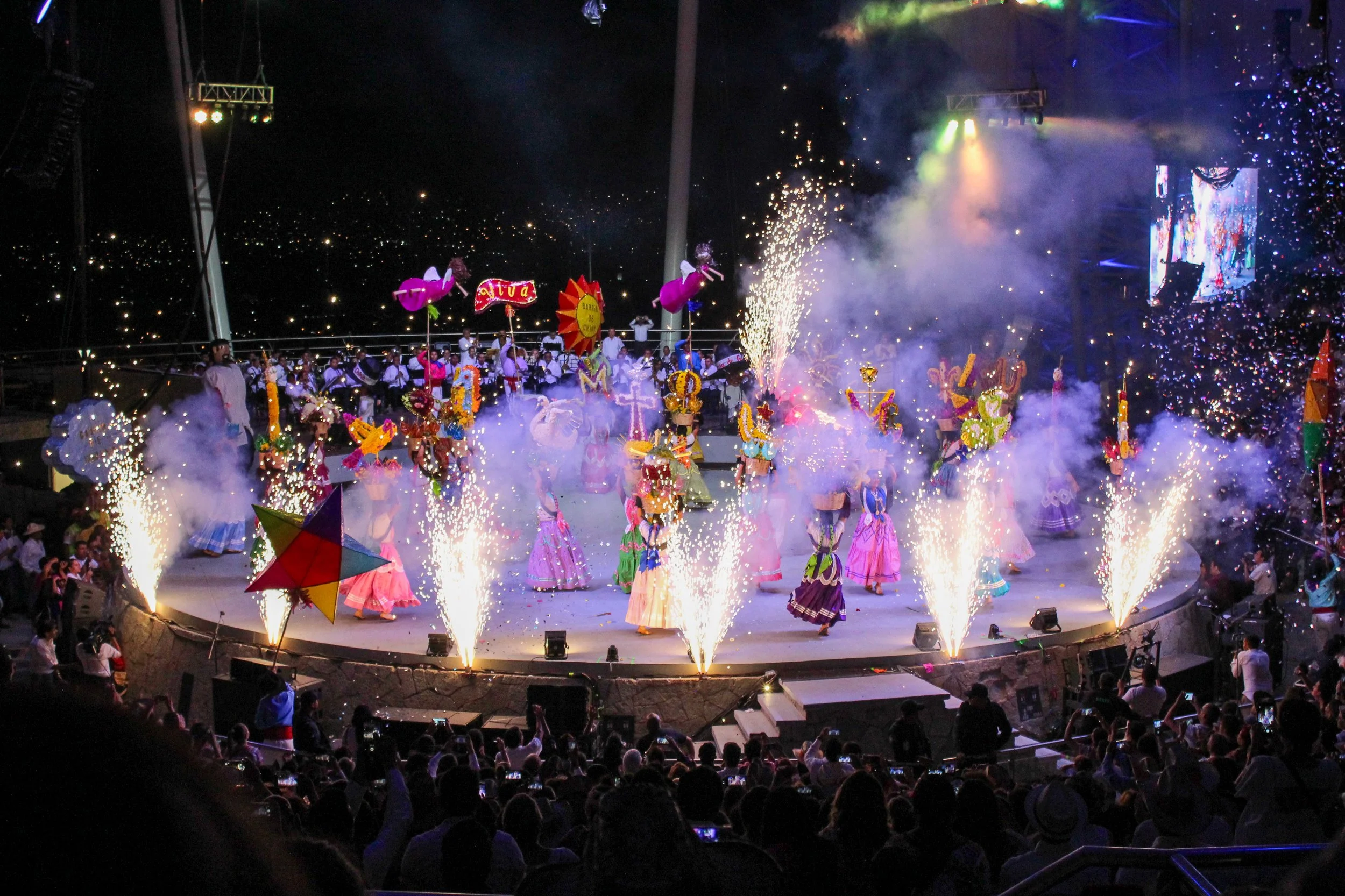 Presentación de baile tradicional con fuegos artificiales y decoraciones coloridas en un escenario circular con público observando.