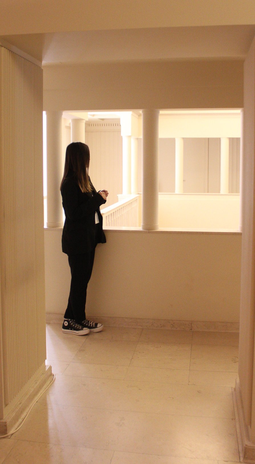 Persona de espaldas con ropa negra y Converse de pie en pasillo con barandilla y columnas blancas.