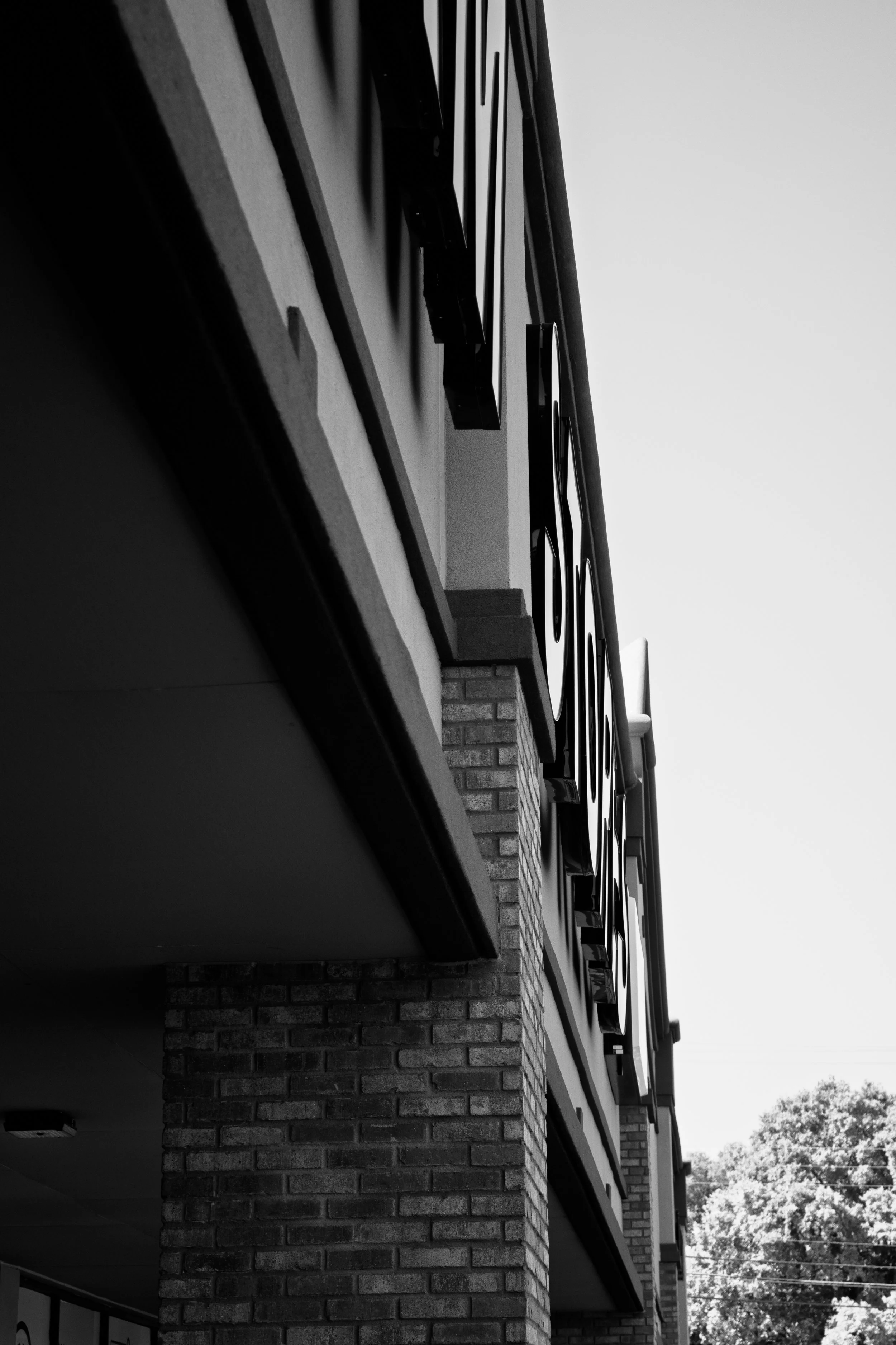 Black and white photo of a building's exterior showing a brick column, balcony with decorative metal railings, and large windows, with trees and a clear sky in the background.