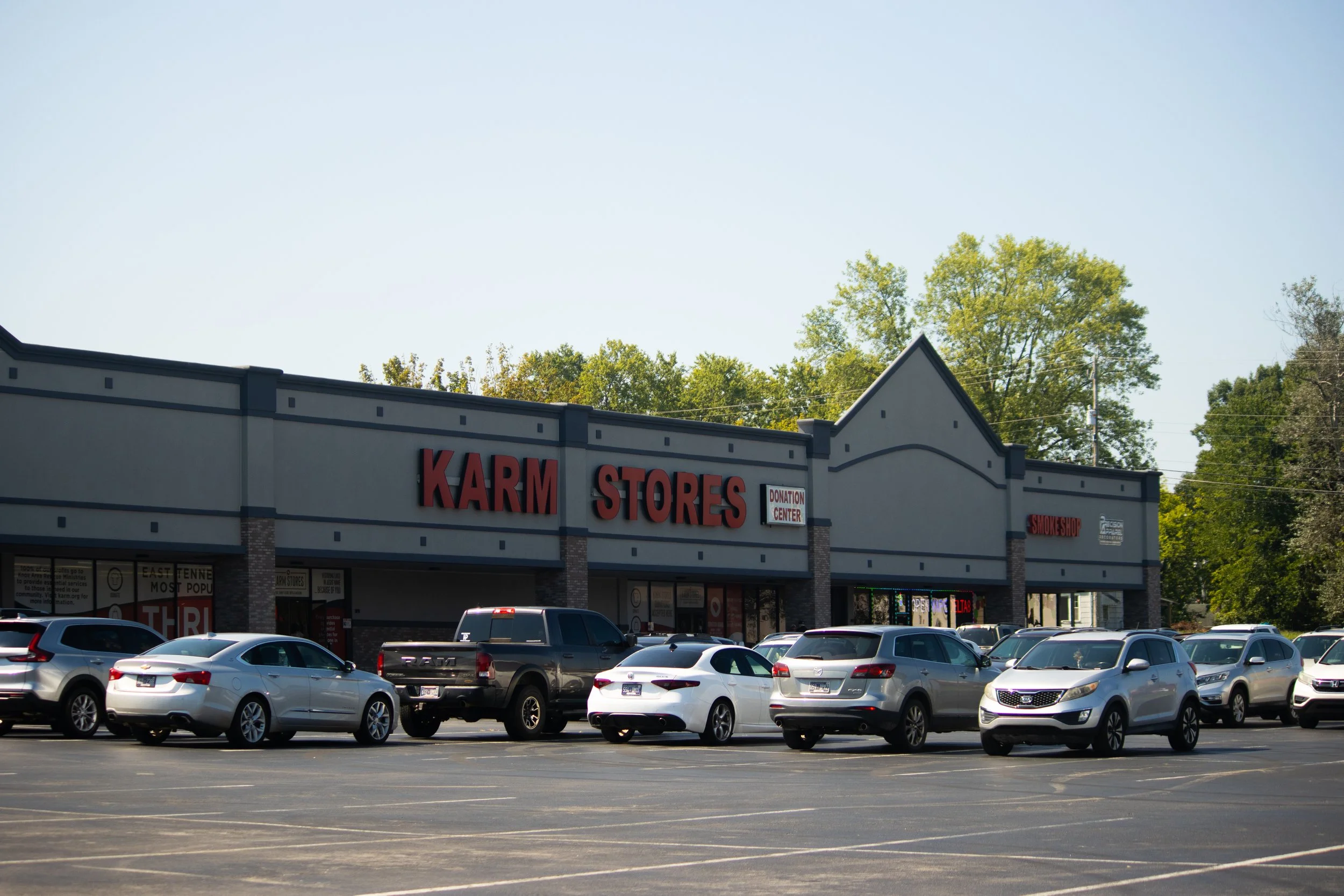 A shopping plaza parking lot with several cars parked in front of a store called 'KARM STORES' and a donation center sign visible, surrounded by trees and clear sky.