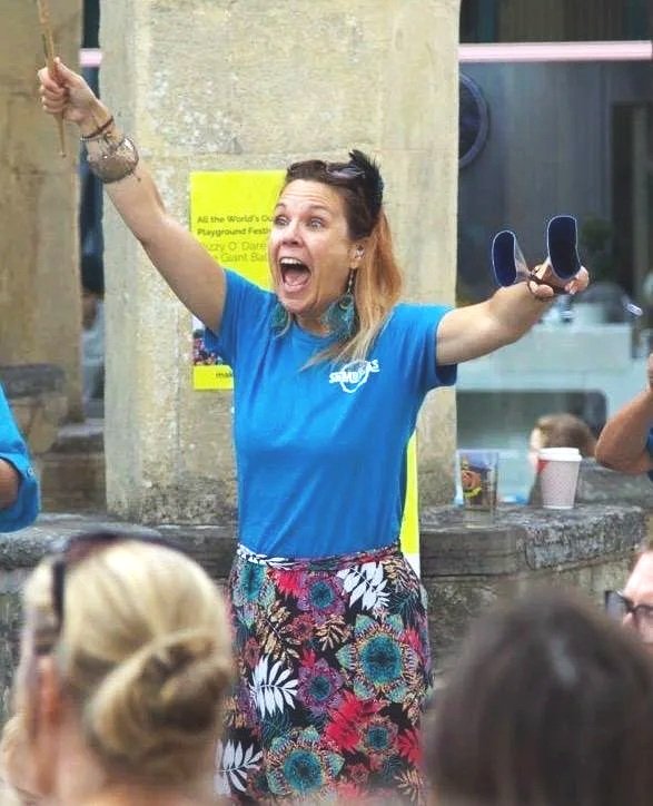 A woman with light skin and reddish hair, wearing a blue shirt and floral skirt, is cheering with her arms raised and holding sunglasses in one hand, in front of a concrete pillar, at an outdoor event with people in the crowd.