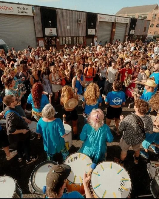 Large crowd gathered outside a building for a protest or demonstration, with people holding signs and drums.