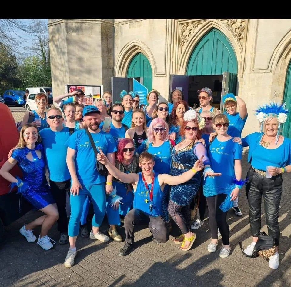 Group of people dressed in blue costumes and accessories, posing outside a building with gothic-style doors, celebrating a themed event or parade.