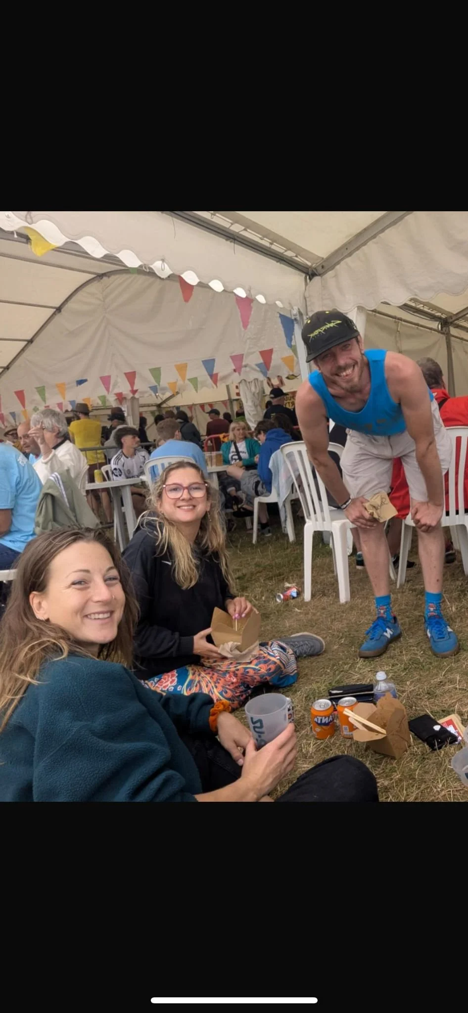 Three people sitting on the grass at a festive outdoor event. They are smiling and enjoying food and drinks. A man is standing, leaning forward, wearing a helmet and athletic clothing. The background shows a crowded tent with colorful bunting and tables with many people.