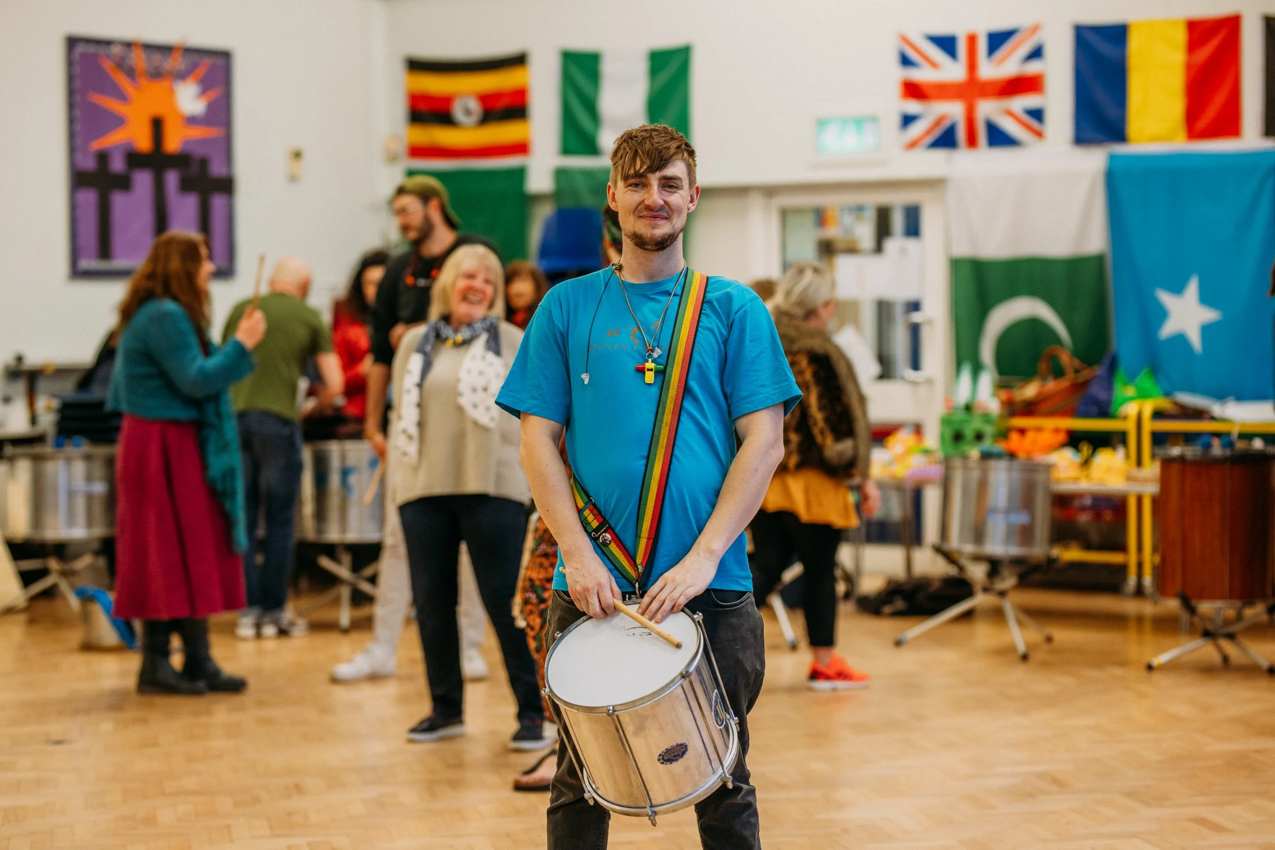 A young man holding a drum and drumsticks, smiling, with colorful rainbow lanyard around his neck, in a community center decorated with international flags and people socializing in the background.