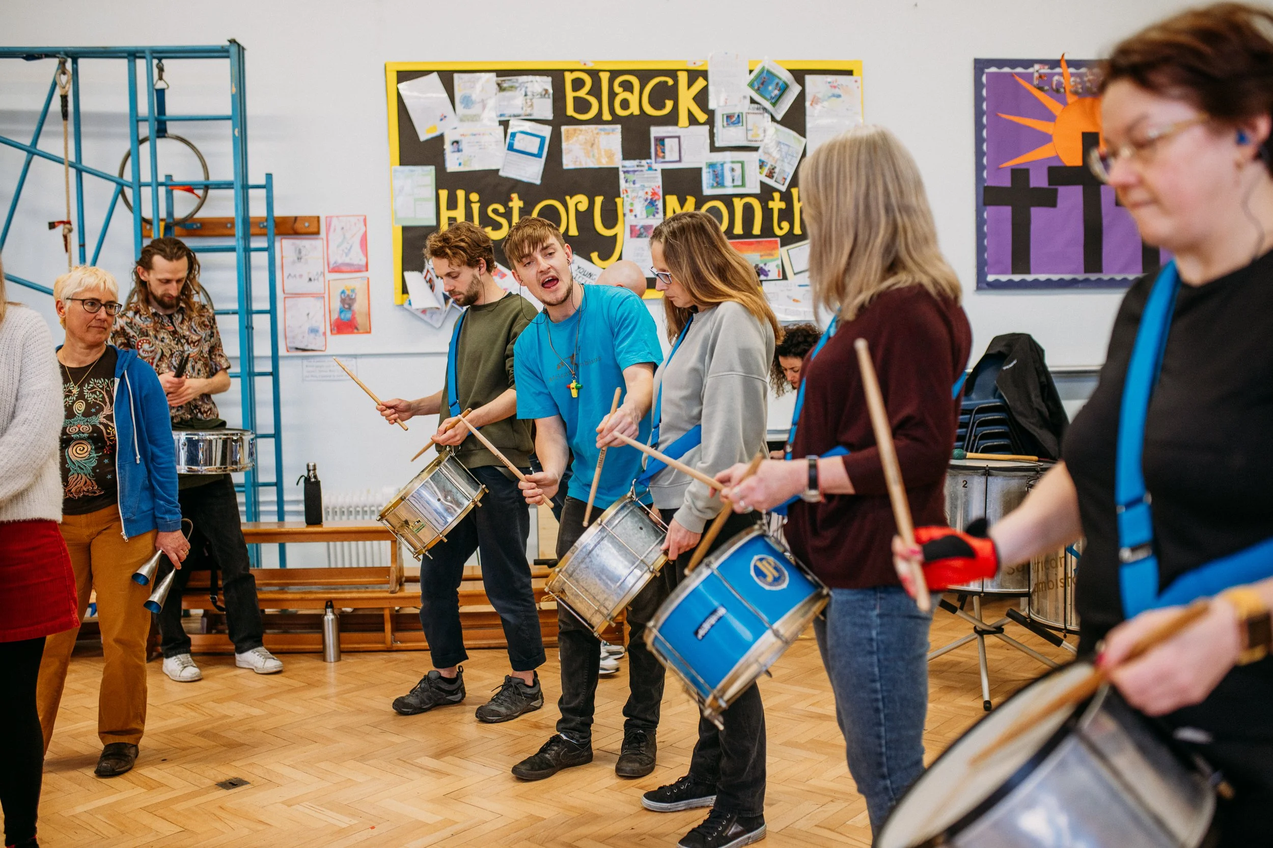 People participating in a drumming circle indoors during Black History Month celebration, with a decorated bulletin board in the background.