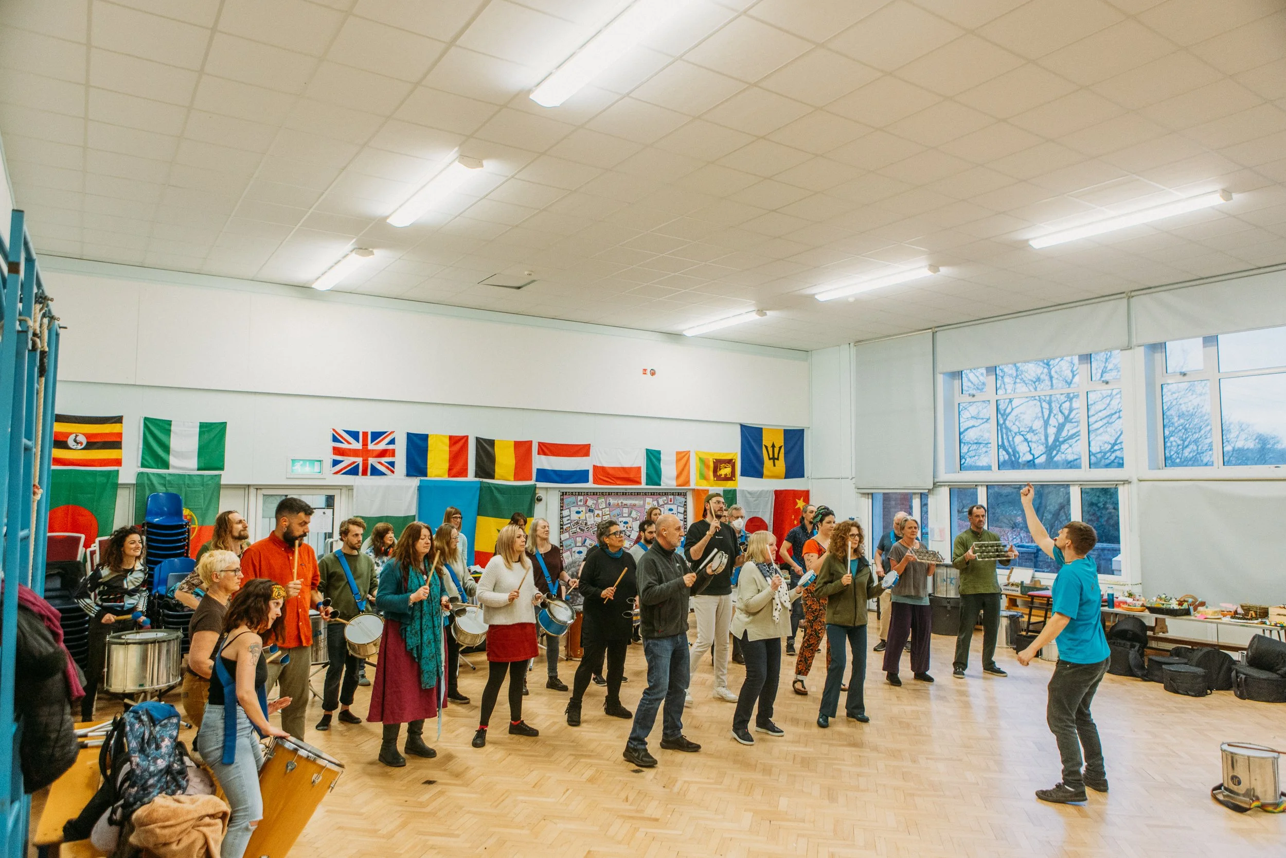 A diverse group of people participating in a music rehearsal or workshop in a spacious room. The group is holding various percussion instruments such as drums and shakers, led by a conductor or instructor at the front. The room has large windows, colorful flags from different countries hanging on the walls, and scattered chairs and bags.