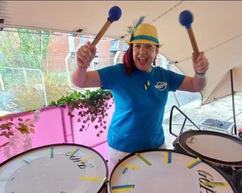 A woman wearing a yellow hat with a feather, a blue t-shirt, and white pants, holding drumsticks and smiling at the camera behind a set of drums. There is a pink backdrop and outdoor setting in the background.
