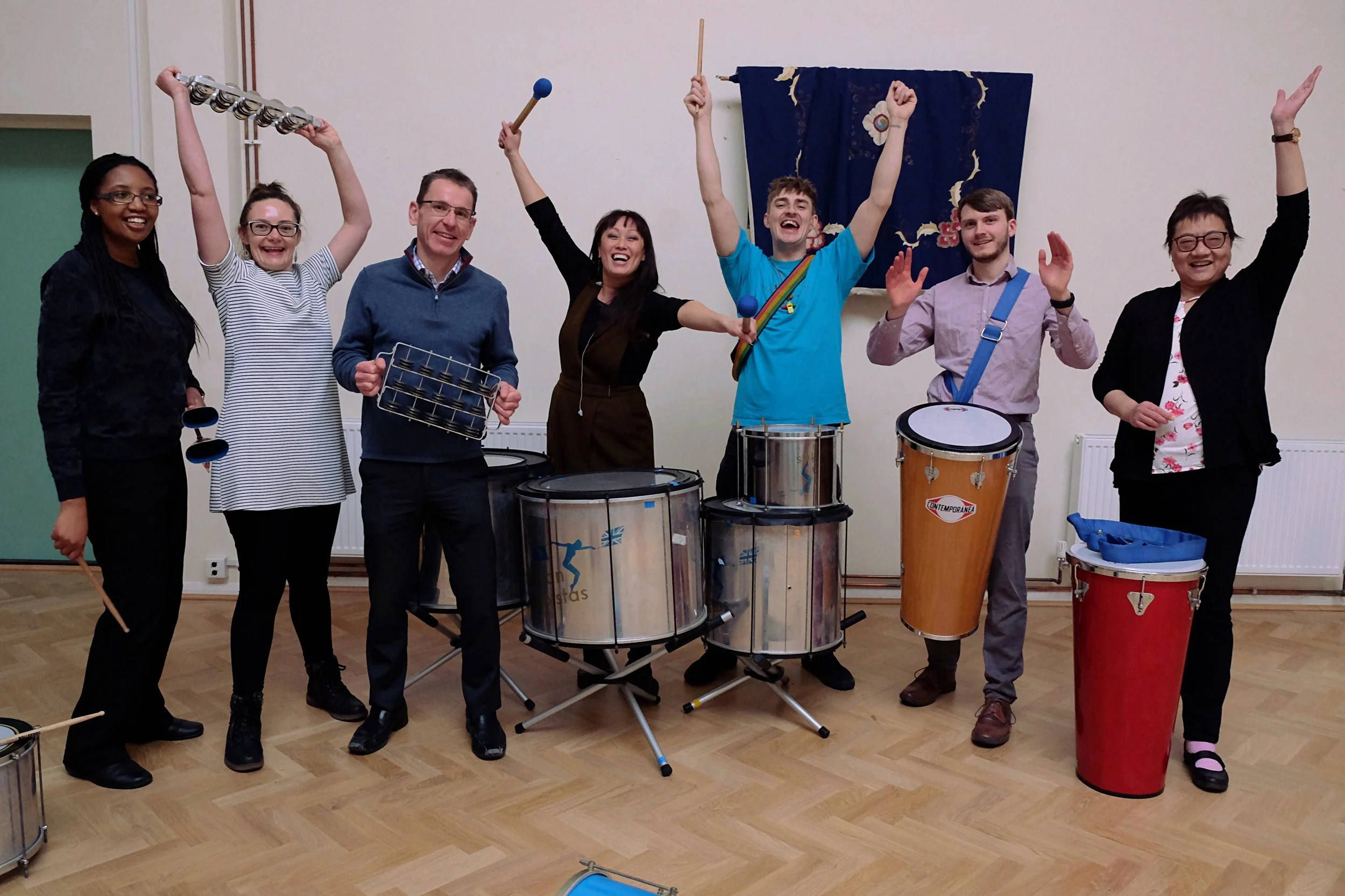 Group of eight people smiling and holding percussion instruments in a room with wooden floors and a decorative wall hanging.