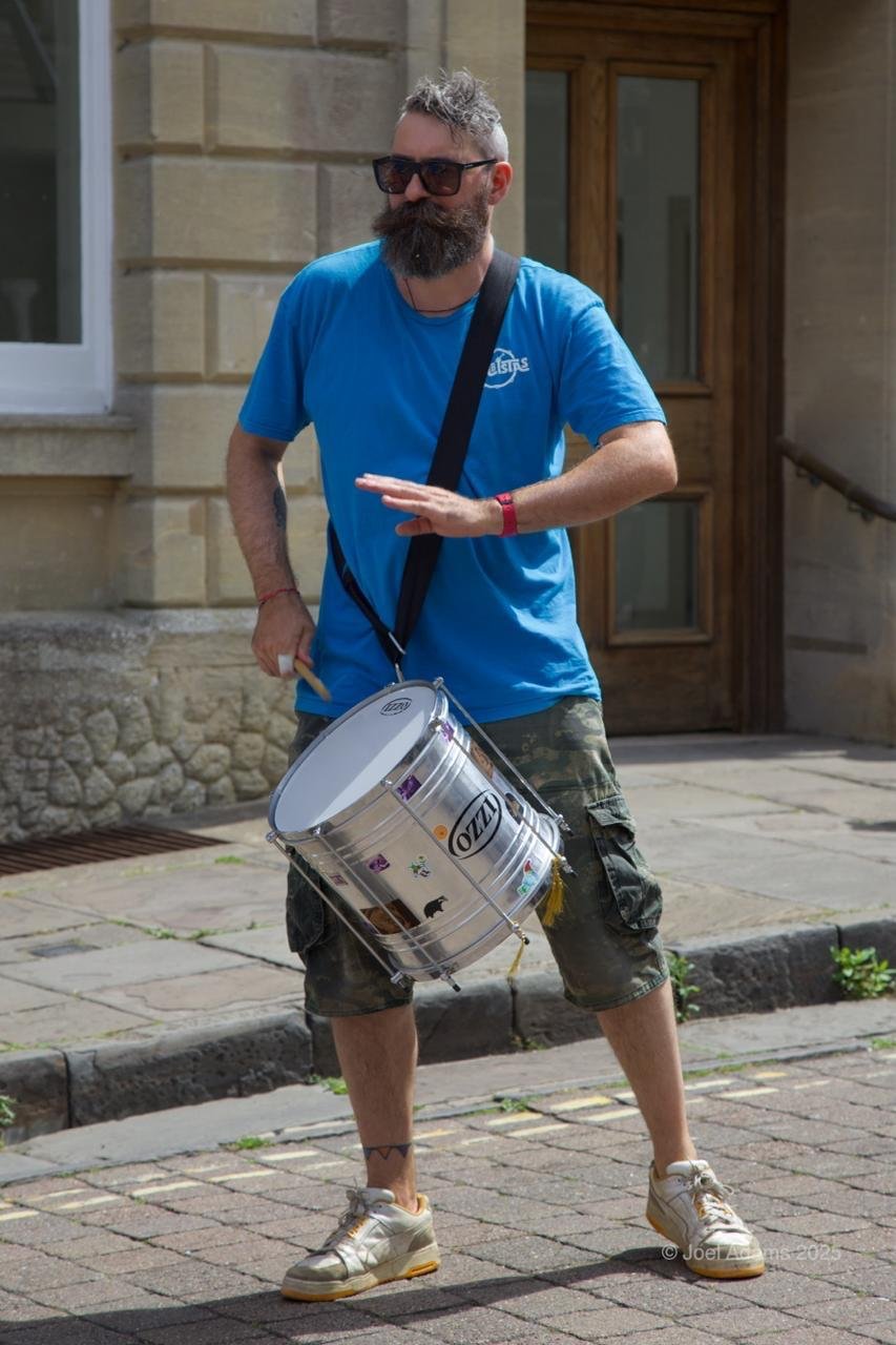 A man with a beard and sunglasses wearing a blue T-shirt, camo shorts, and sneakers, standing on a brick sidewalk playing a snare drum attached to his waist, outside a building with stone and wooden doors.
