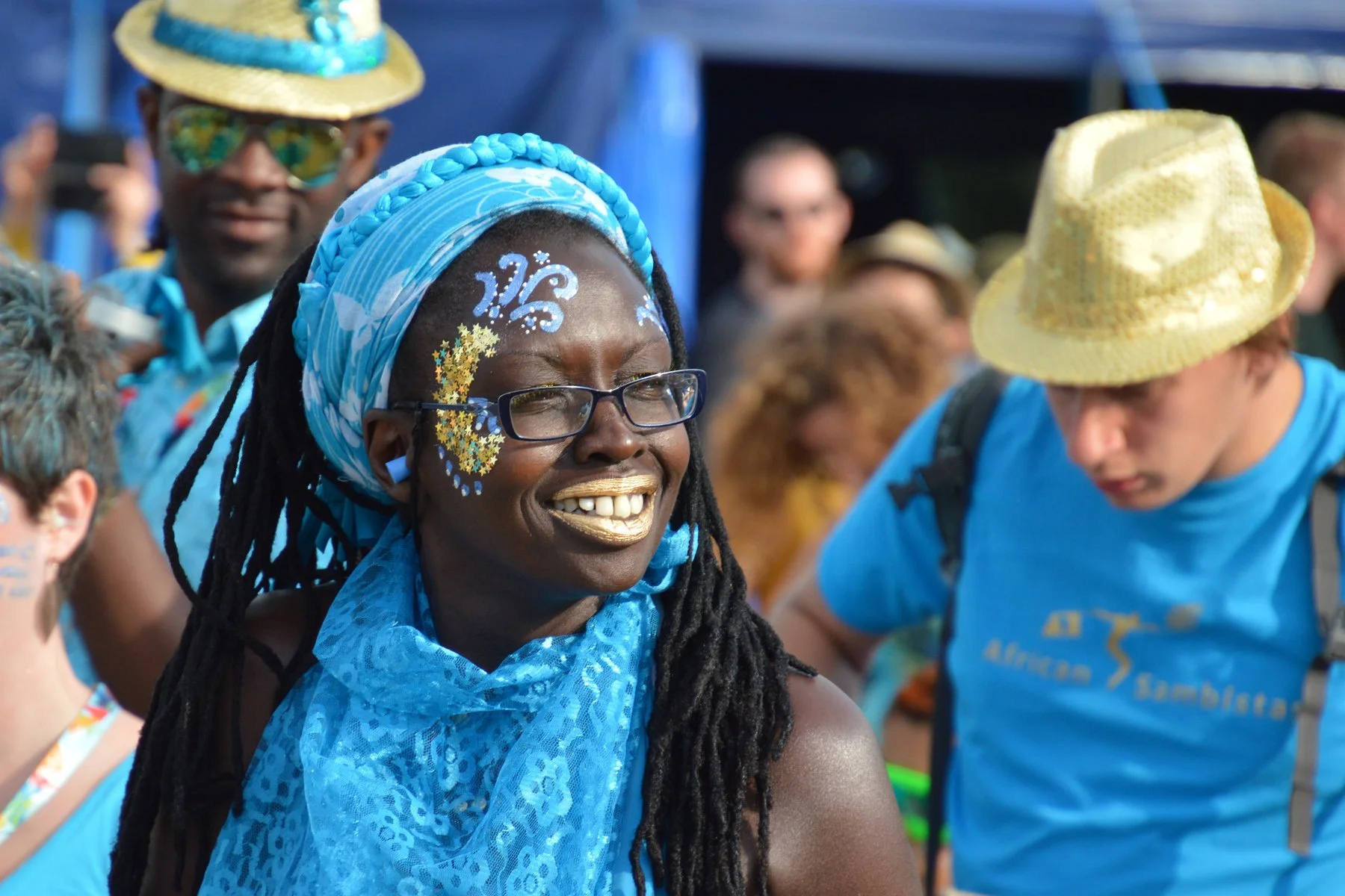 A group of people outdoors, dressed in blue, participating in a festive event. The woman in the foreground has face paint and gold lipstick, is wearing glasses, a blue headwrap, and a blue scarf. She is smiling, and others around her are wearing hats and casual clothing.