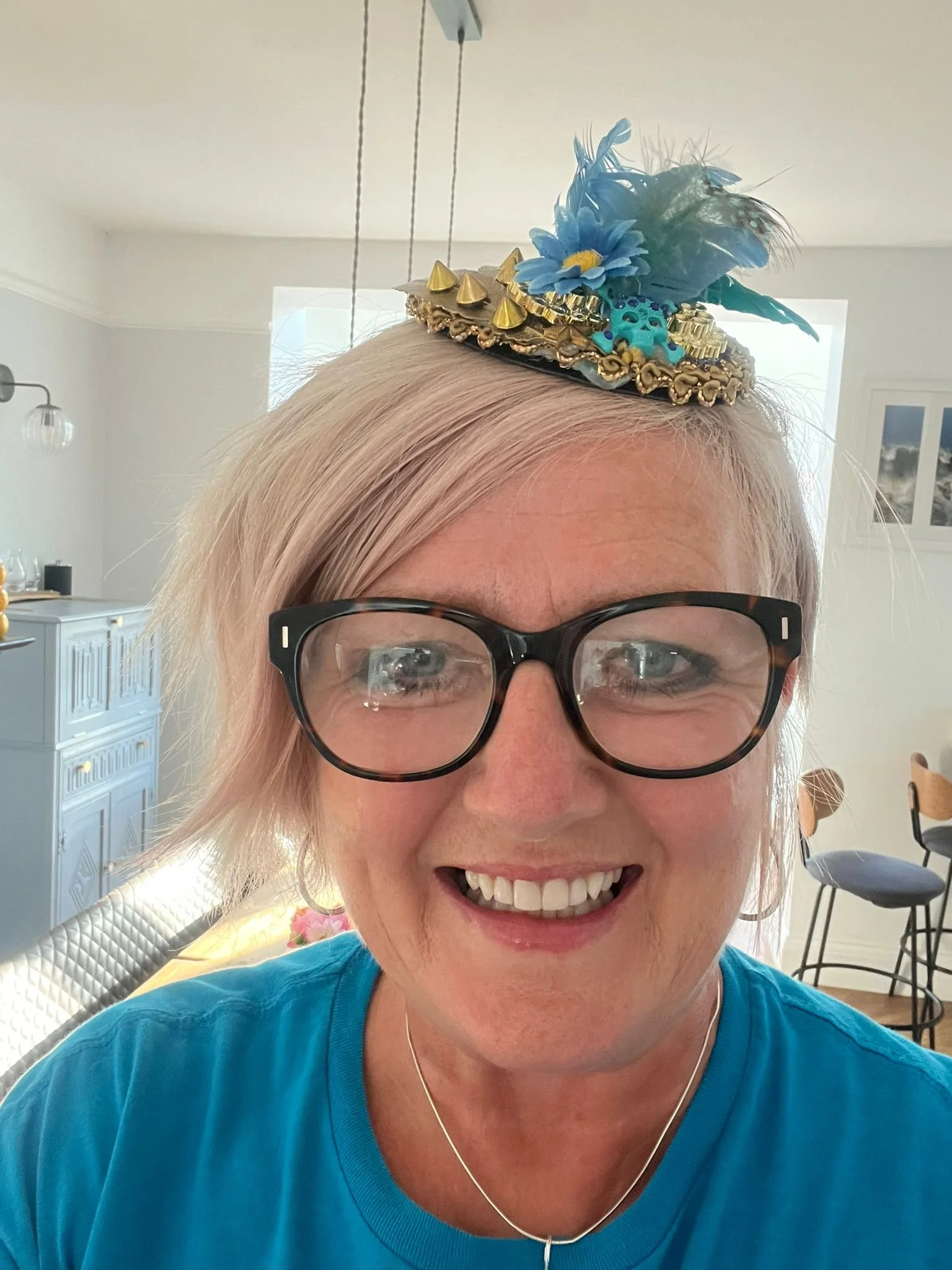 A woman smiling with glasses, wearing a teal shirt, and a festive fascinator hat decorated with blue feathers, yellow and turquoise flowers, gold spikes, and lace, in a bright room with light-colored walls, a blue dresser, and a few chairs in the background.