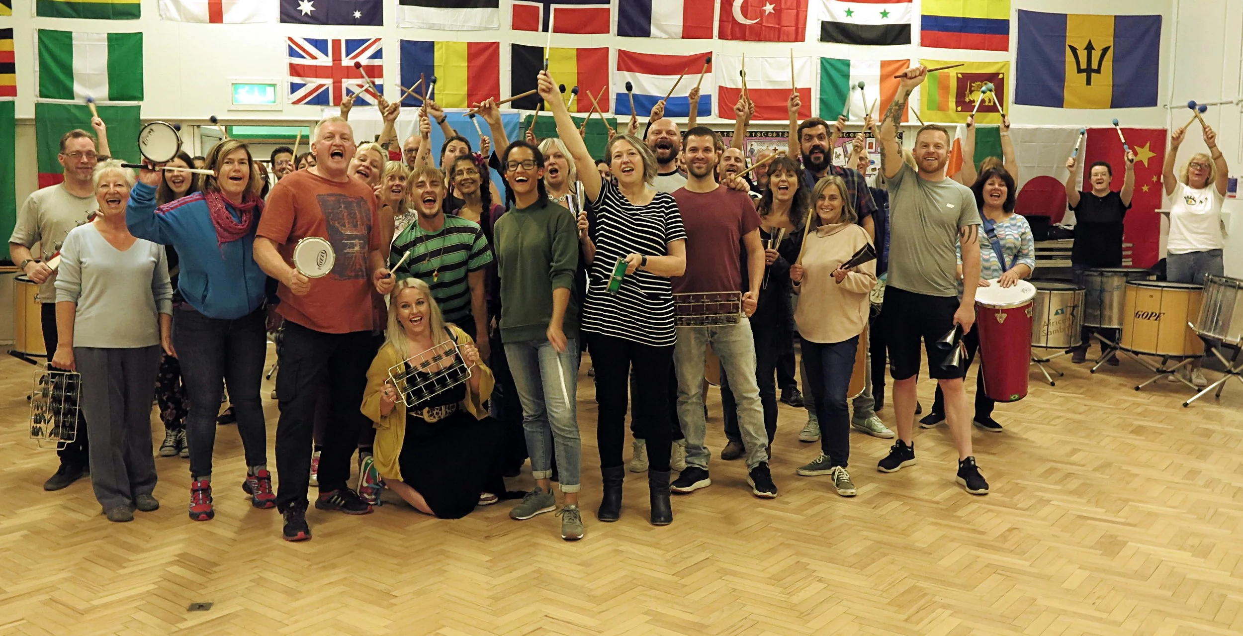 A large group of people smiling and holding musical instruments in a room decorated with international flags, suggesting a music or cultural event.