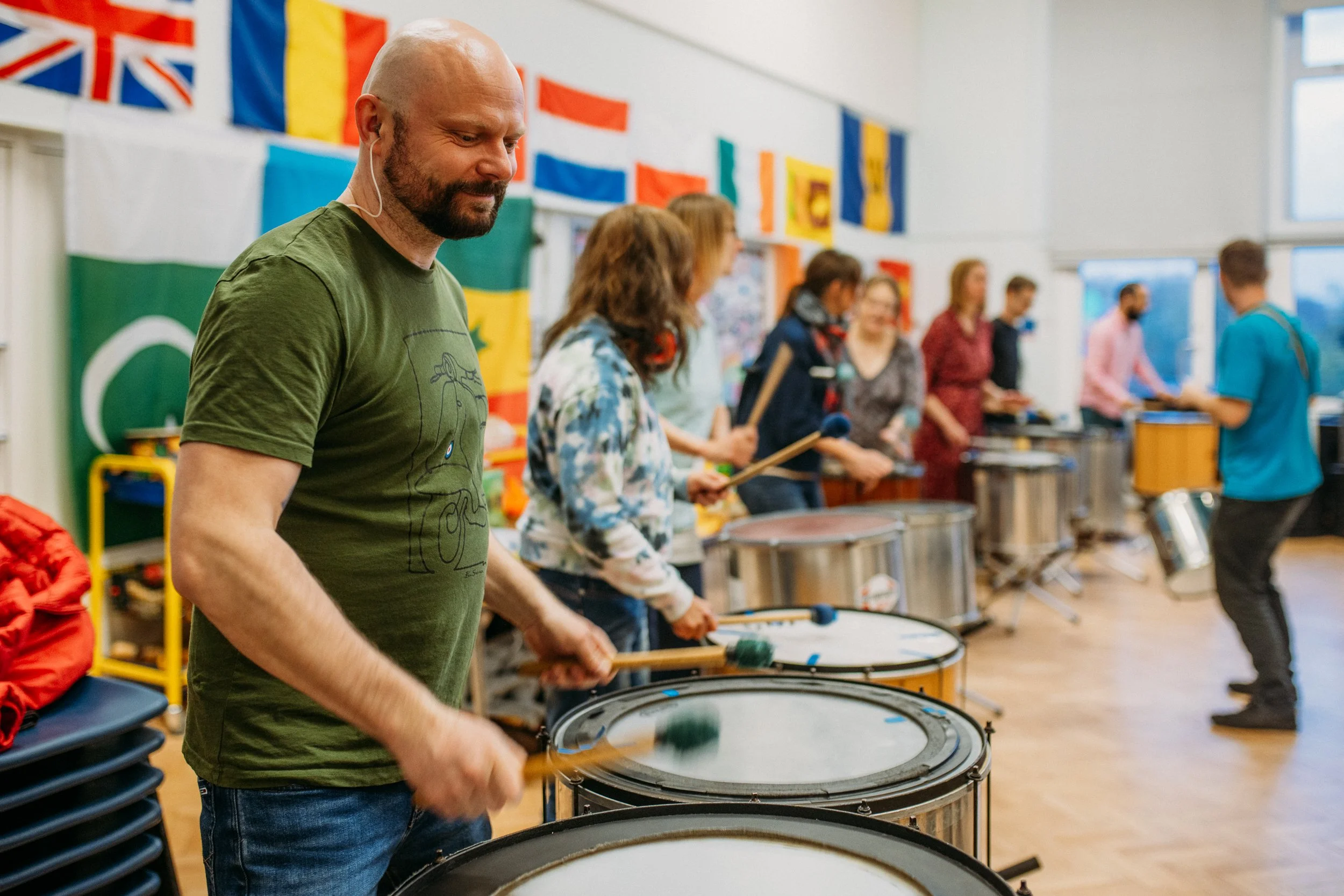 A group of people playing percussion instruments, lined up in a classroom decorated with international flags on the wall.