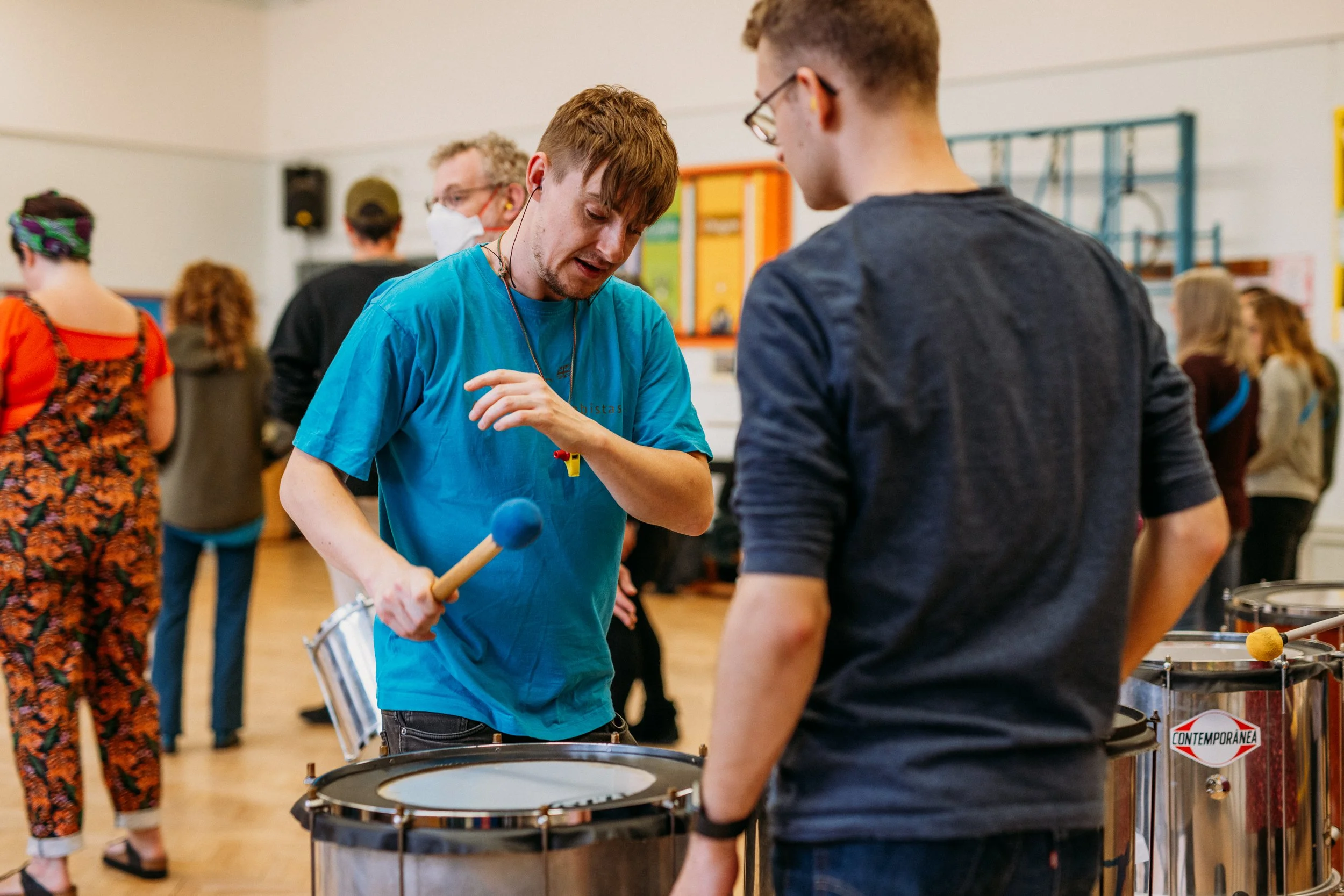 Two young men in a music room, one playing a drum and the other talking, with a group of people in the background.