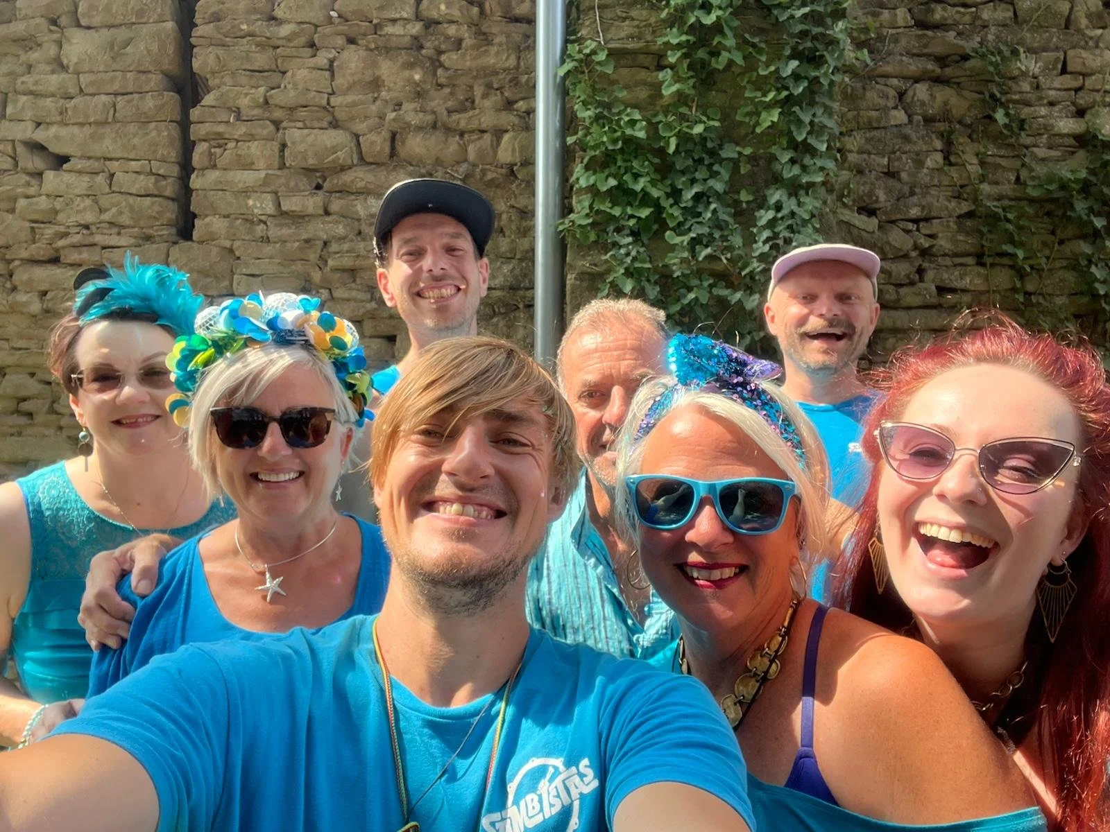 A group of nine people smiling and taking a selfie outdoors in front of a stone wall with ivy. They are wearing colorful and festive clothing, sunglasses, and accessories.