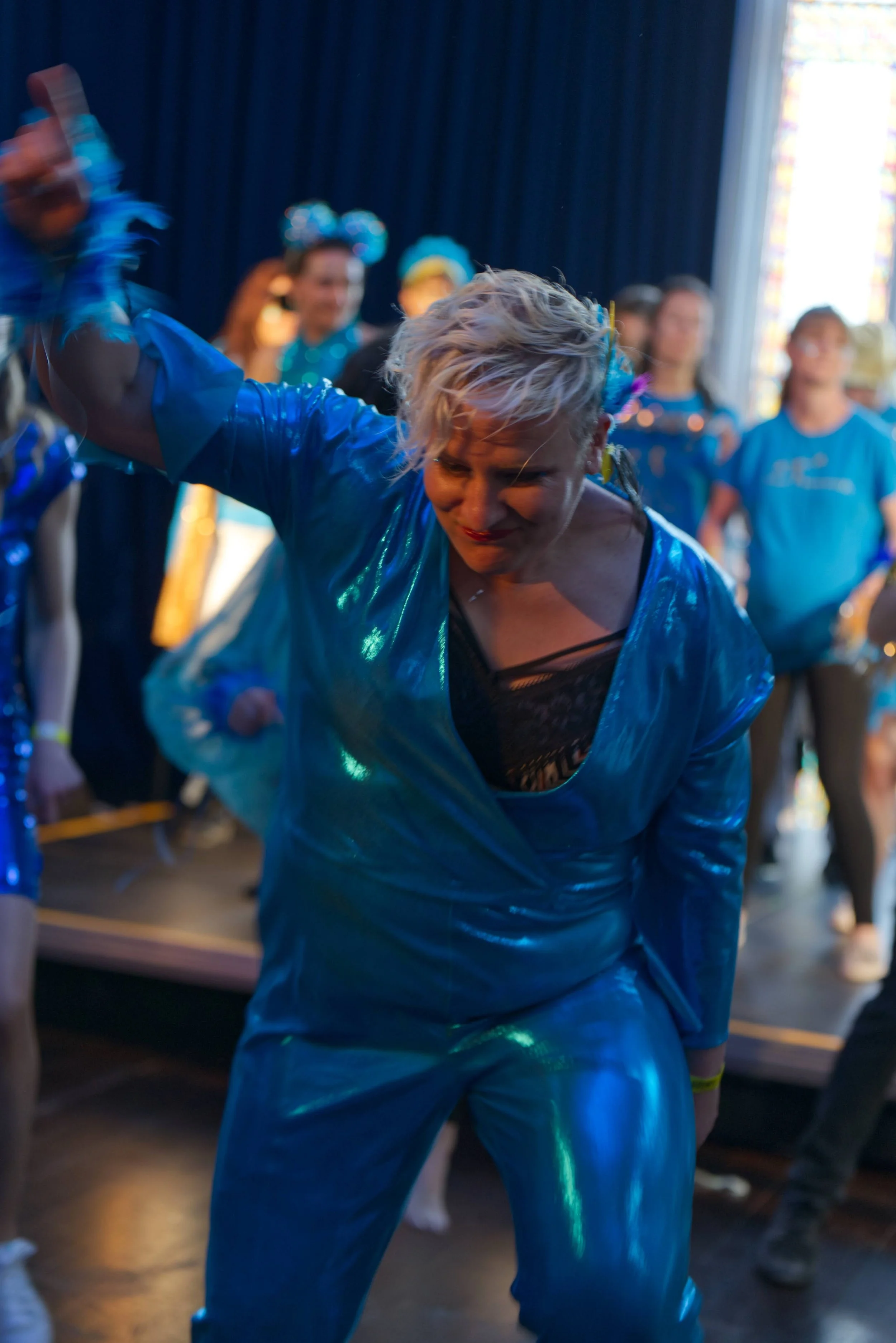 Person with short blonde hair dancing or performing in a shiny blue outfit, surrounded by other people in colorful costumes, in a lively indoor setting.