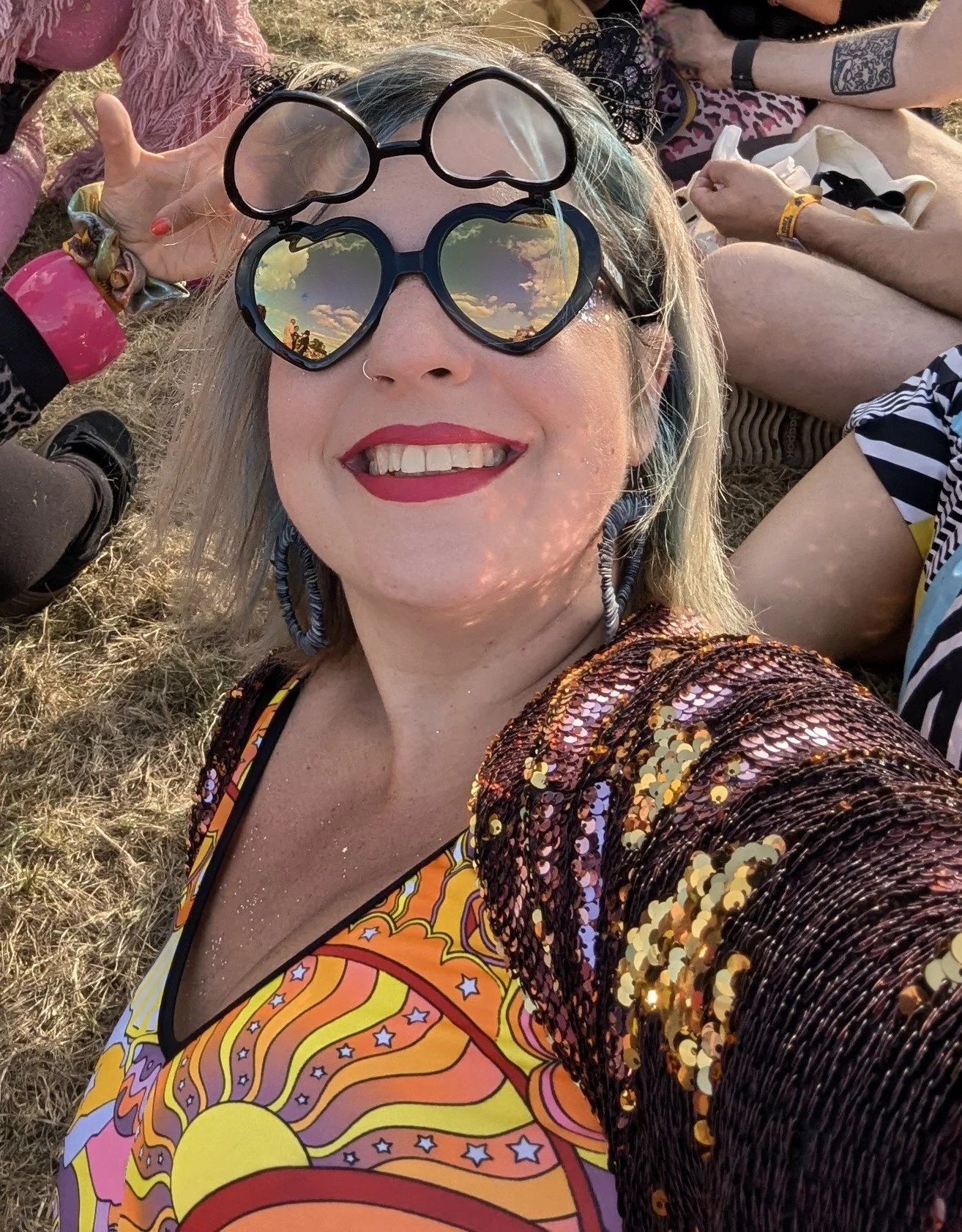 A woman taking a selfie at an outdoor event, smiling, wearing heart-shaped sunglasses reflecting a sunset sky, a colorful psychedelic dress, and a sequined jacket. People sitting on the ground are visible in the background.