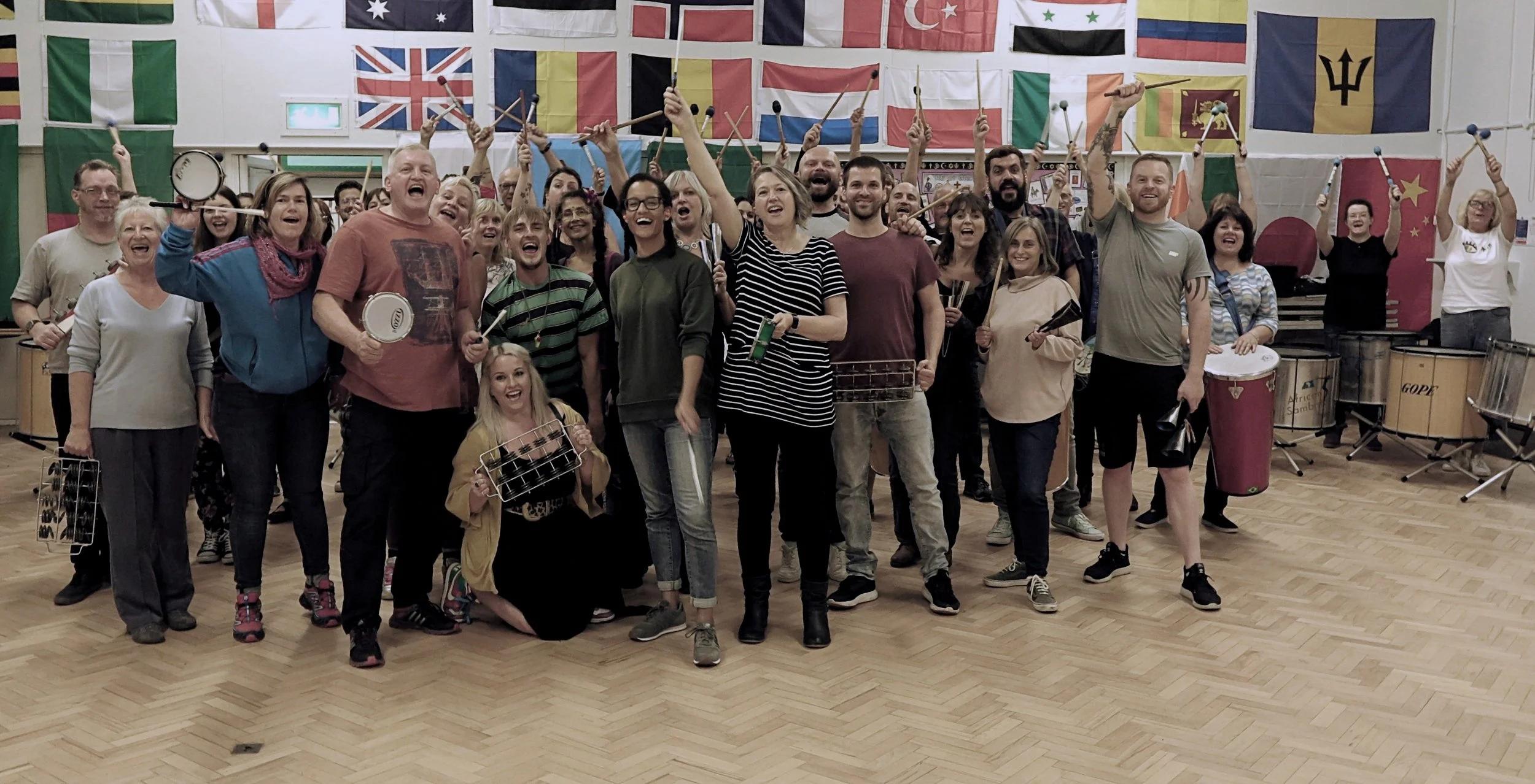 A large group of people posing together indoors with musical instruments, surrounded by international flags hanging from the ceiling. Samba Band. Community