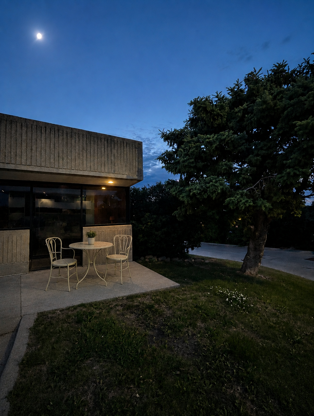 An outdoor patio area at dusk with a small white table and two white chairs, a potted plant on the table, a building with large windows and a textured concrete exterior, a large tree, and a bright moon in the evening sky.