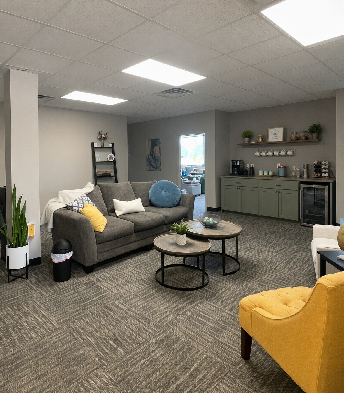 A cozy lounge area with a grey sofa, yellow and white accent pillows, and a blue round pillow. Two round coffee tables with plants and a bowl of decorative objects. A yellow upholstered armchair to the right. A small kitchenette with green cabinets, a coffee machine, and shelves with decor and mugs. A doorway leads to another room with seating. The space has a neutral color palette with earthy tones and modern decor.