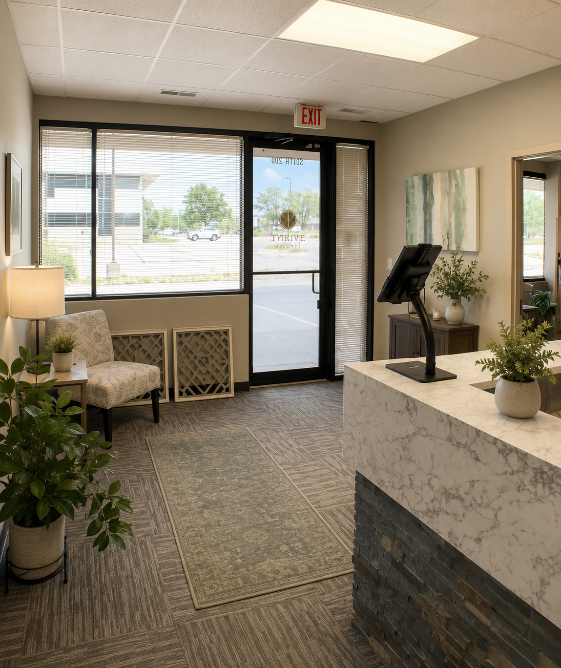Interior of a wellness or medical office reception area with a marble-topped desk, potted plants, wall art, a chair near the window, and a glass door leading outside.
