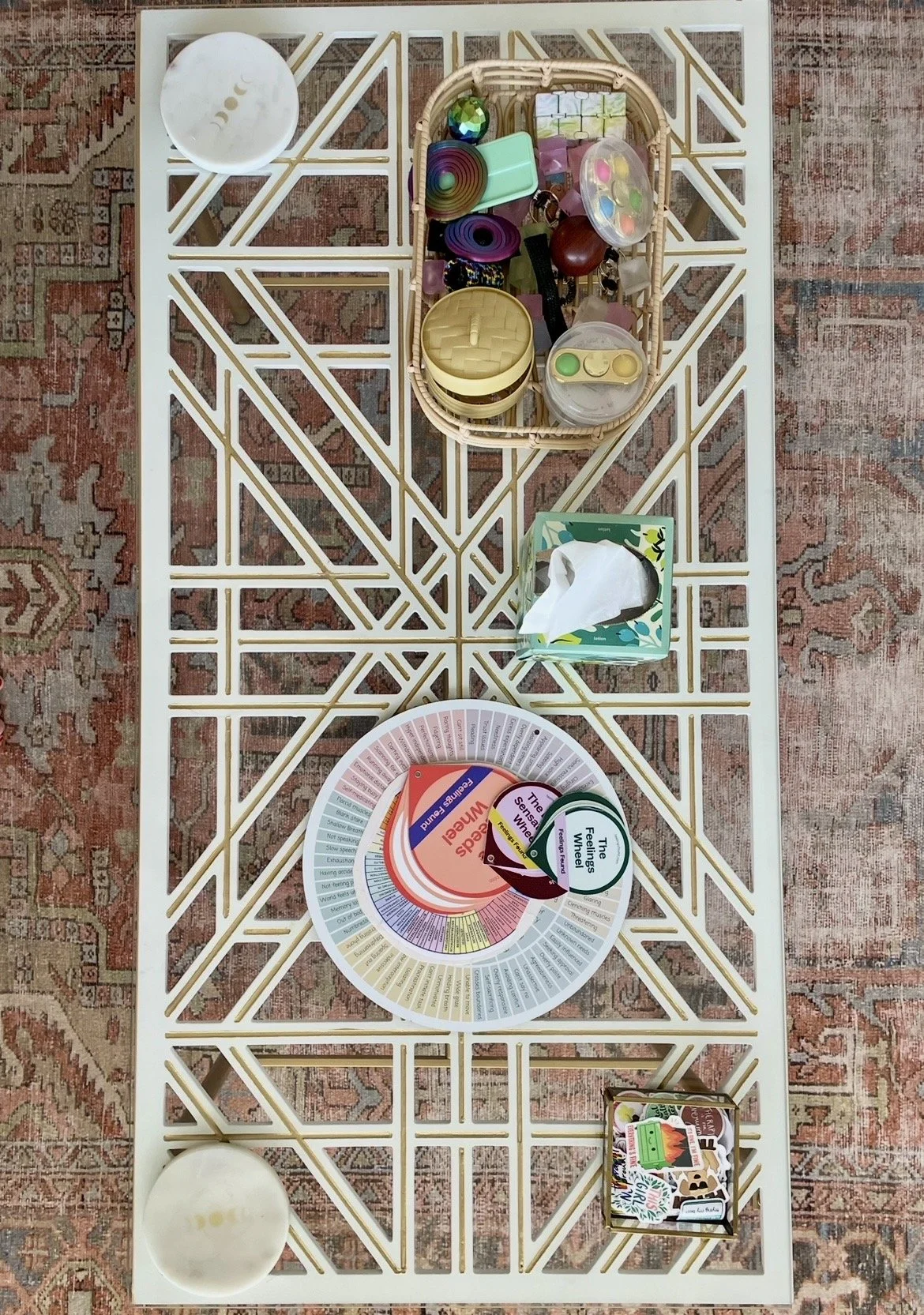 A table with a decorative open lattice design and display items including a large circular color wheel, a deck of colorful playing cards, a tissue box, and various small objects in a basket and on the table. The table is on a patterned rug.