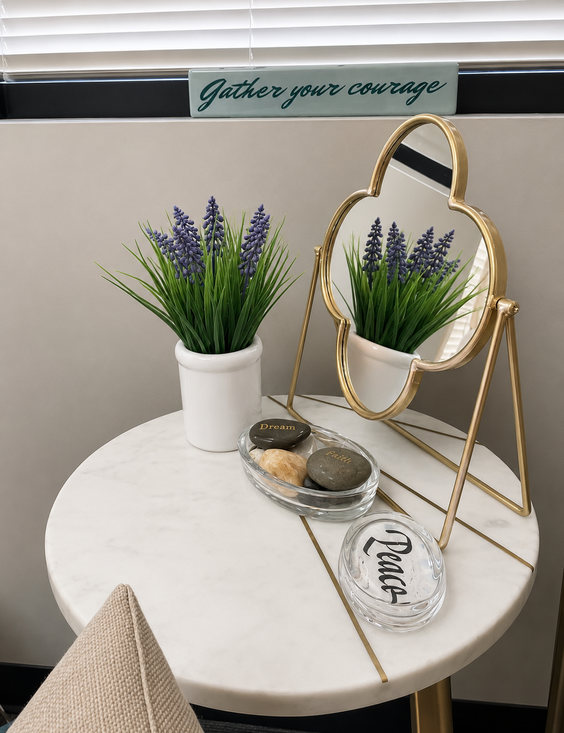 Decorative table with artificial purple flowers in a white pot, a gold-framed mirror, and a small bowl with stones inscribed with 'Dream,' 'Faith,' and 'Peace.' A sign above says 'Gather your courage.'