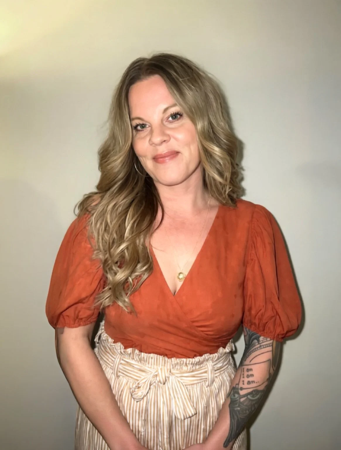 A woman with long, wavy blonde hair wearing an orange top and striped beige pants, standing in front of a plain light-colored wall.