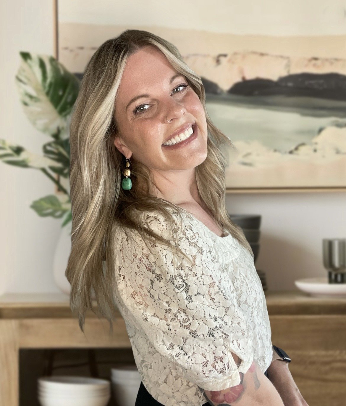 A young woman with blonde hair smiling and posing indoors, wearing a lace top and earrings with green stones, with art and a plant in the background.