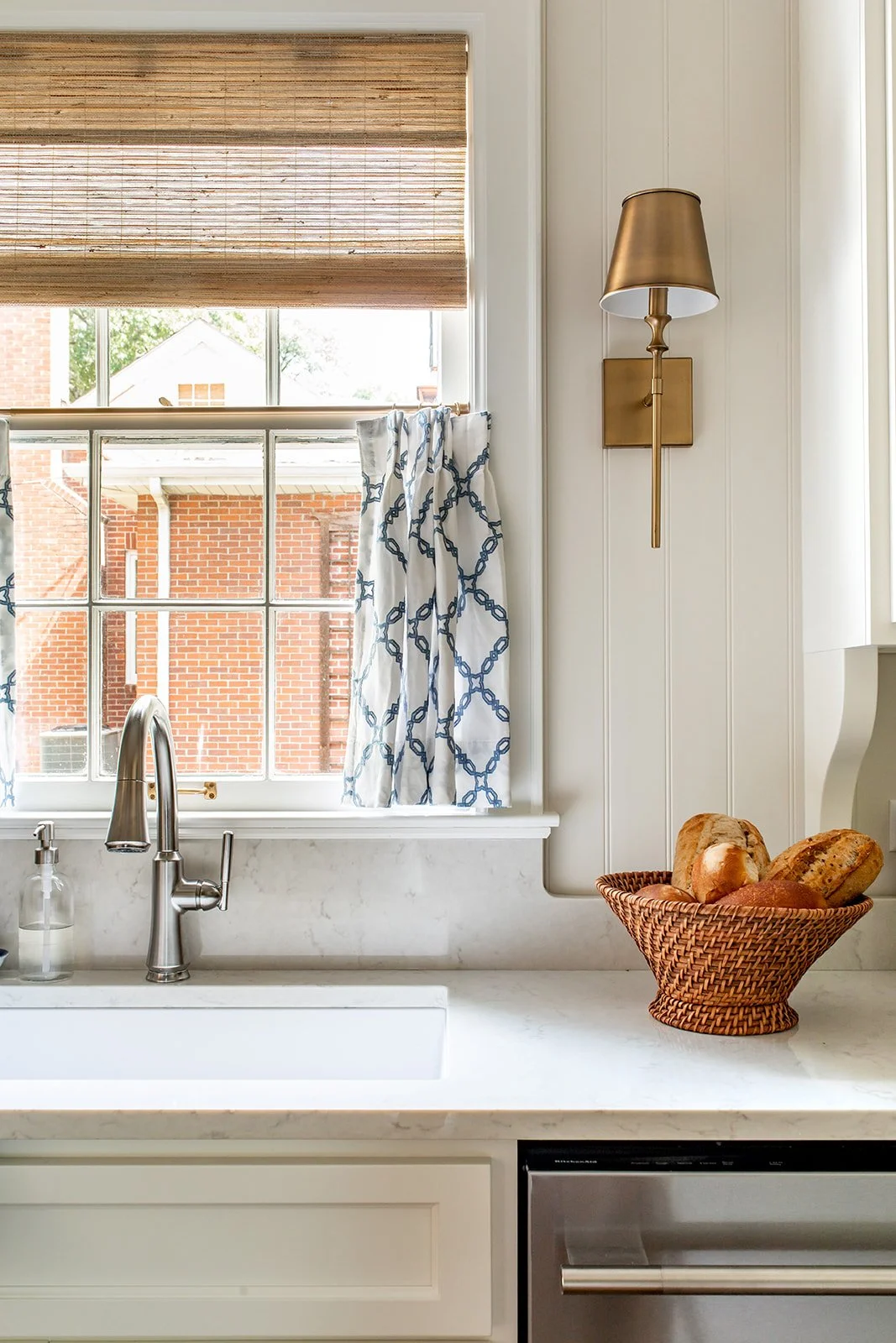 Kitchen sink corner with a brass sconce, marble counter, and blue cafe curtains