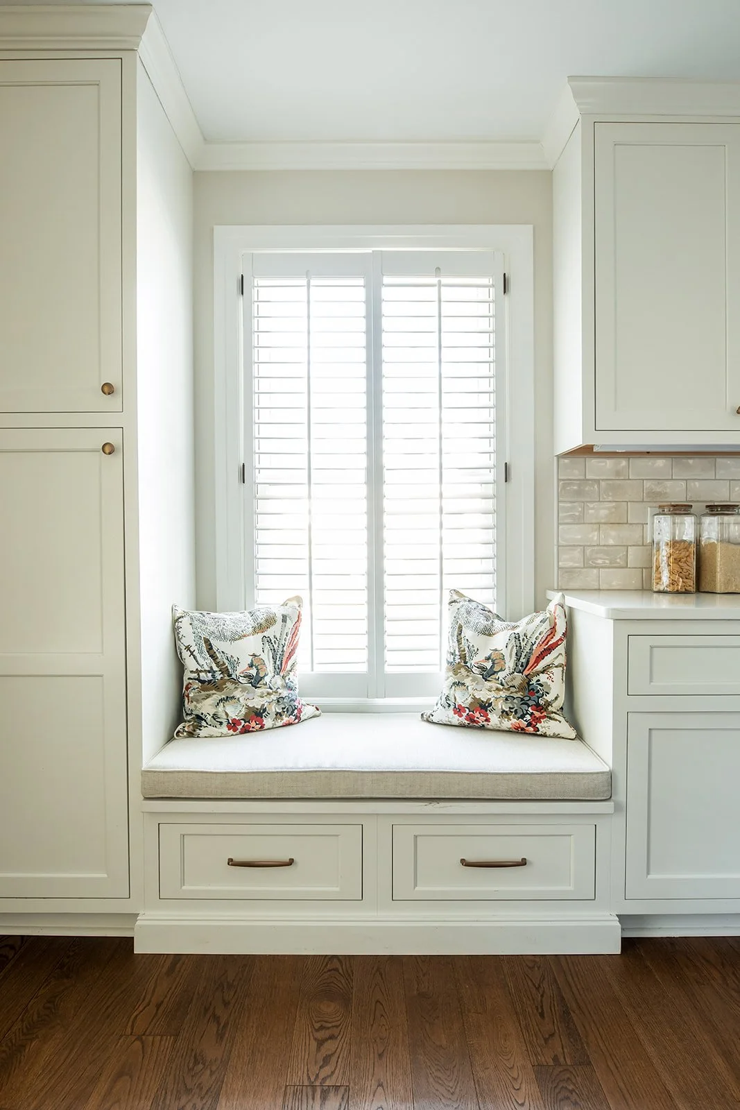 Built-in kitchen window seat with floral pillows beneath plantation shutters
