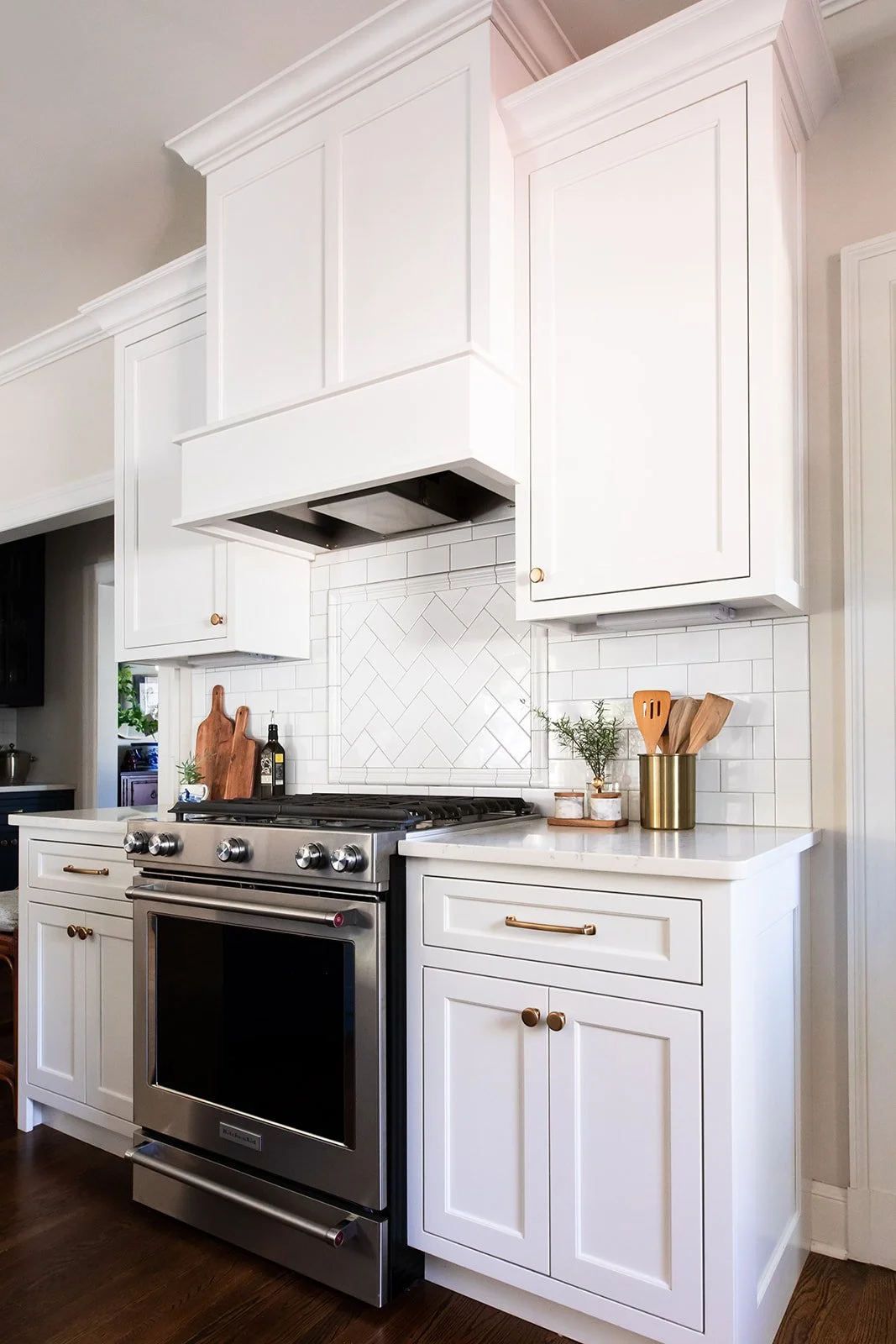 White kitchen range wall with a custom vent hood and herringbone tile backsplash
