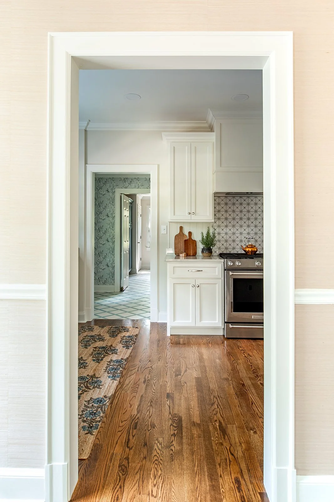 View through doorway into a kitchen with white cabinets, a stove, and wooden cutting boards on the counter, hardwood flooring, and a hallway with patterned wallpaper in the background.