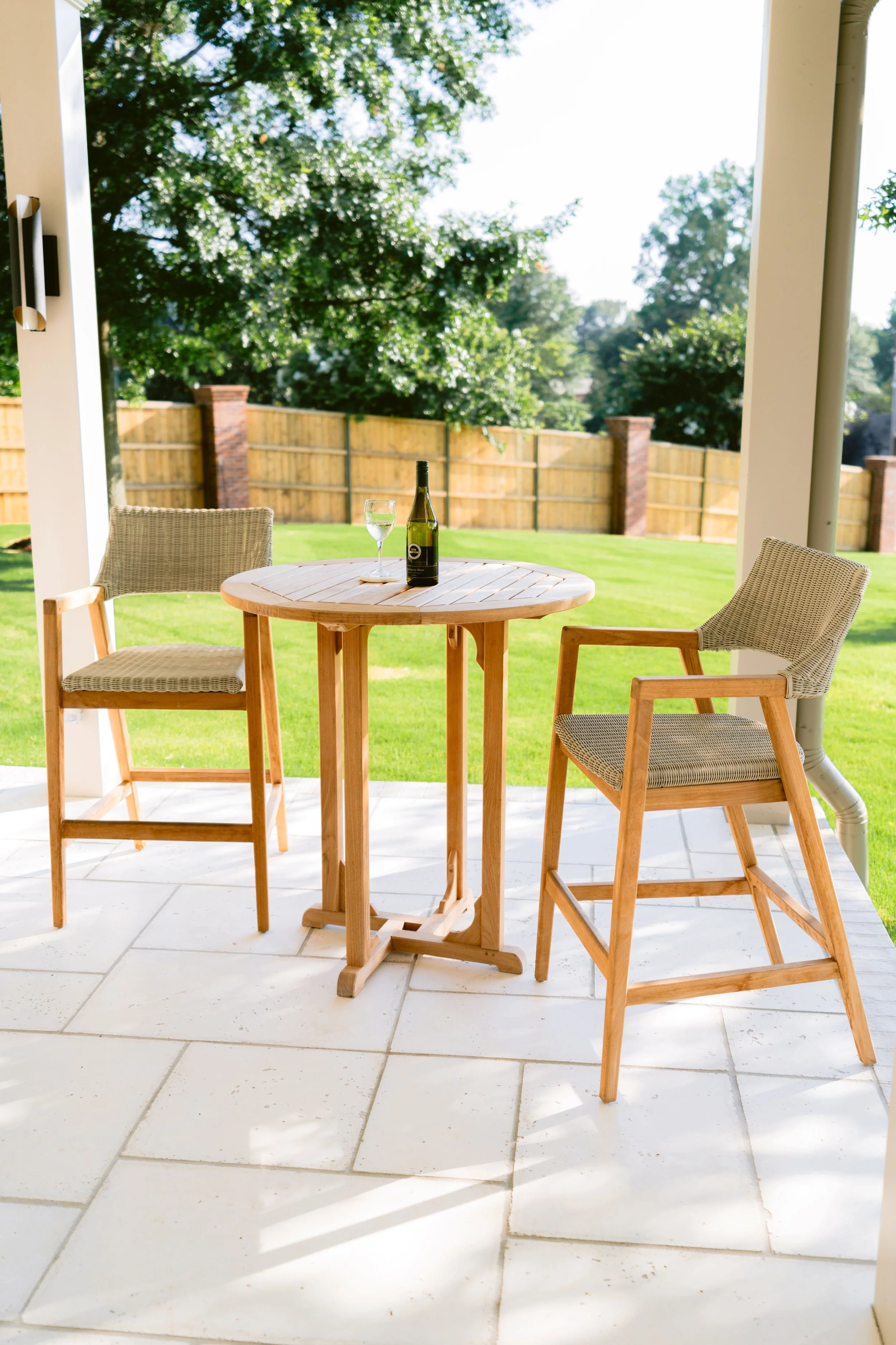 Bar-height outdoor table with two woven stools on a stone patio overlooking the lawn