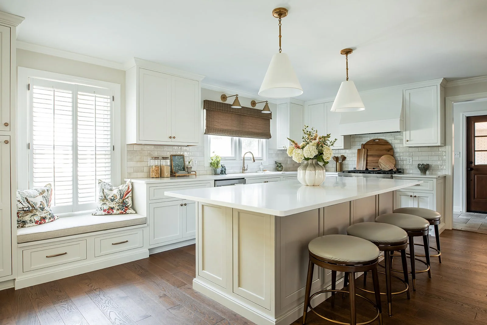 Warm white kitchen with a center island, window seat, brass pendants, and woven shades