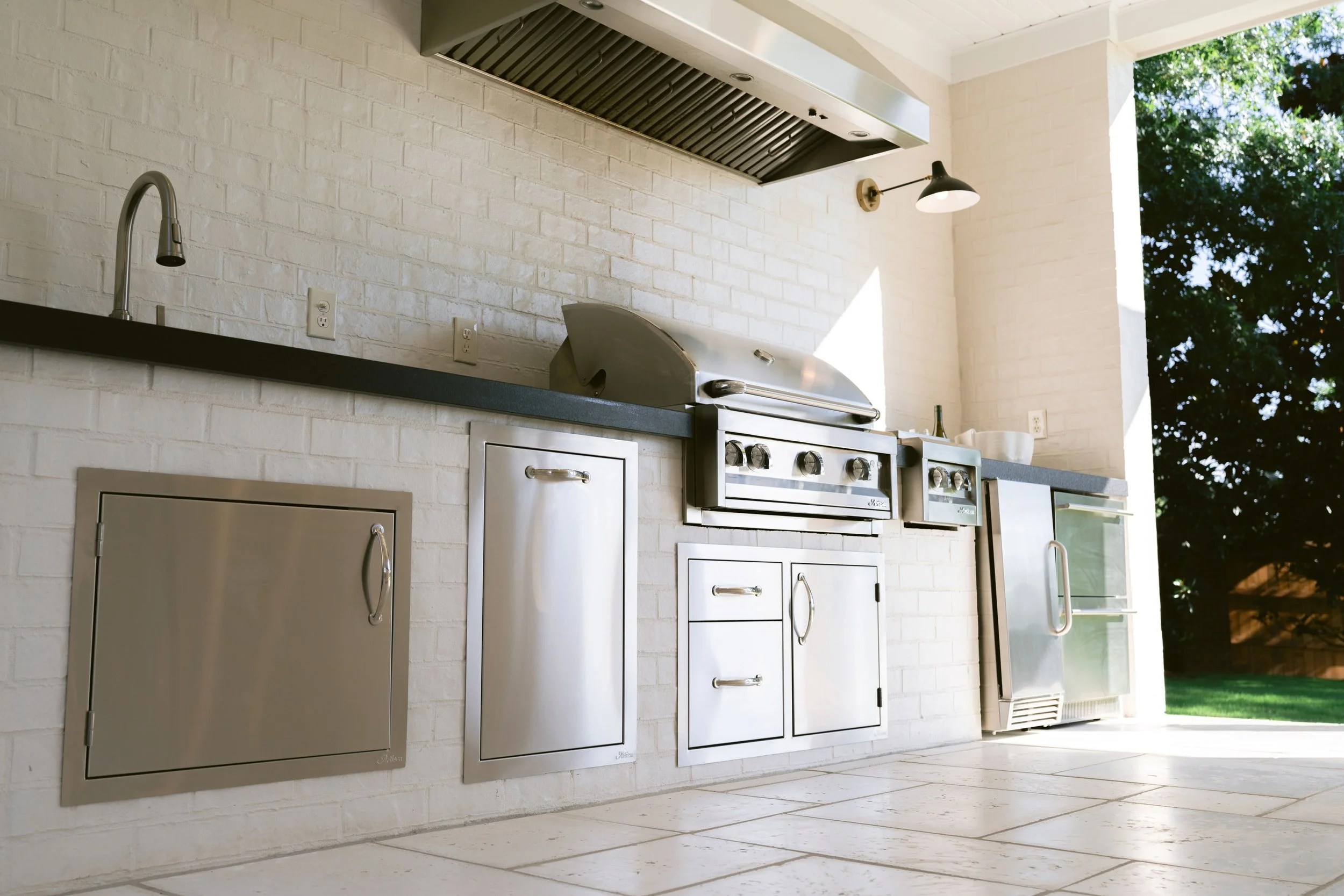 Outdoor kitchen with a stainless grill, vent hood, and mini fridge against white brick