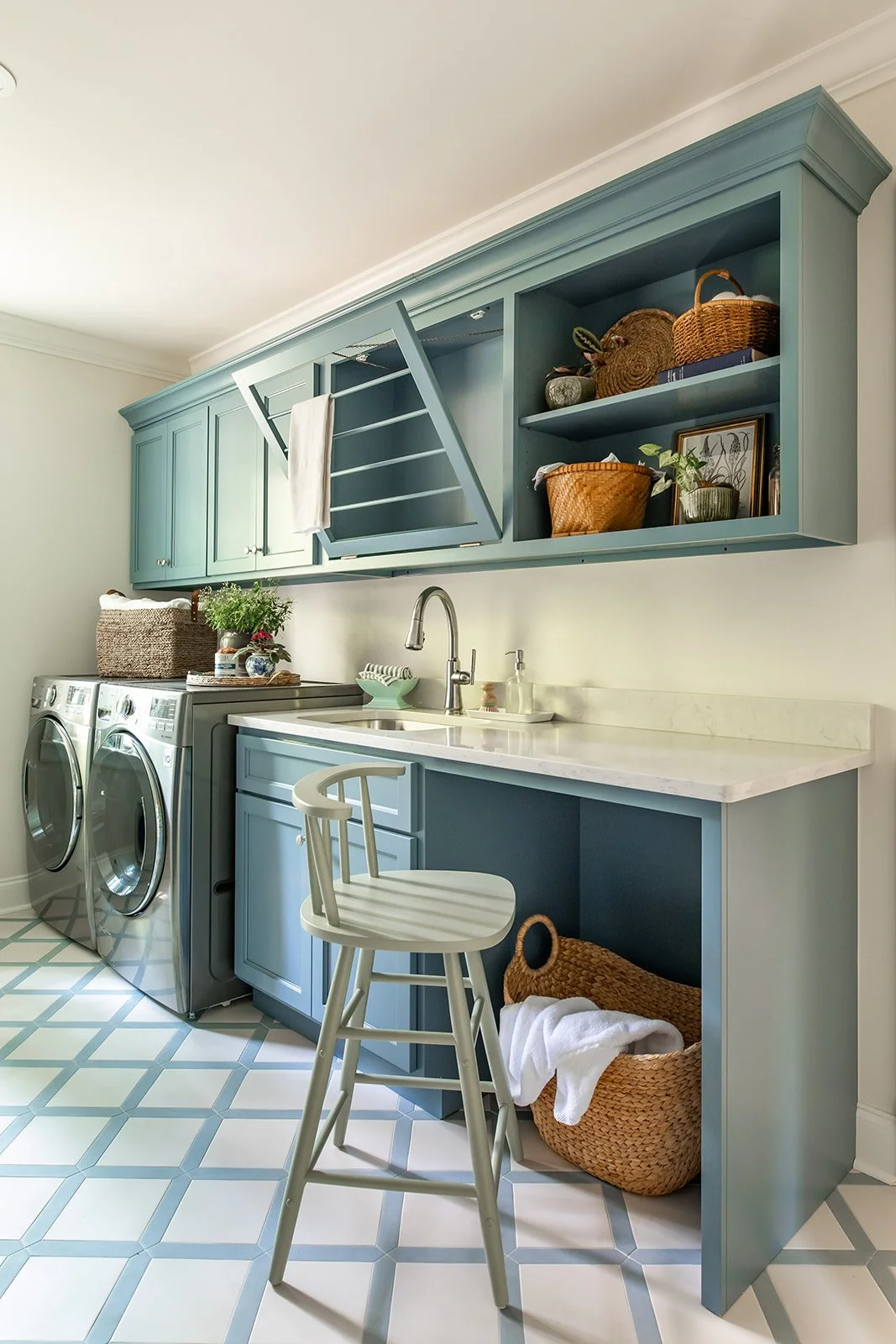 A laundry room with blue cabinets, a white countertop, and a tiled floor. There are two front-loading washing machines, a white chair, and woven baskets for storage.
