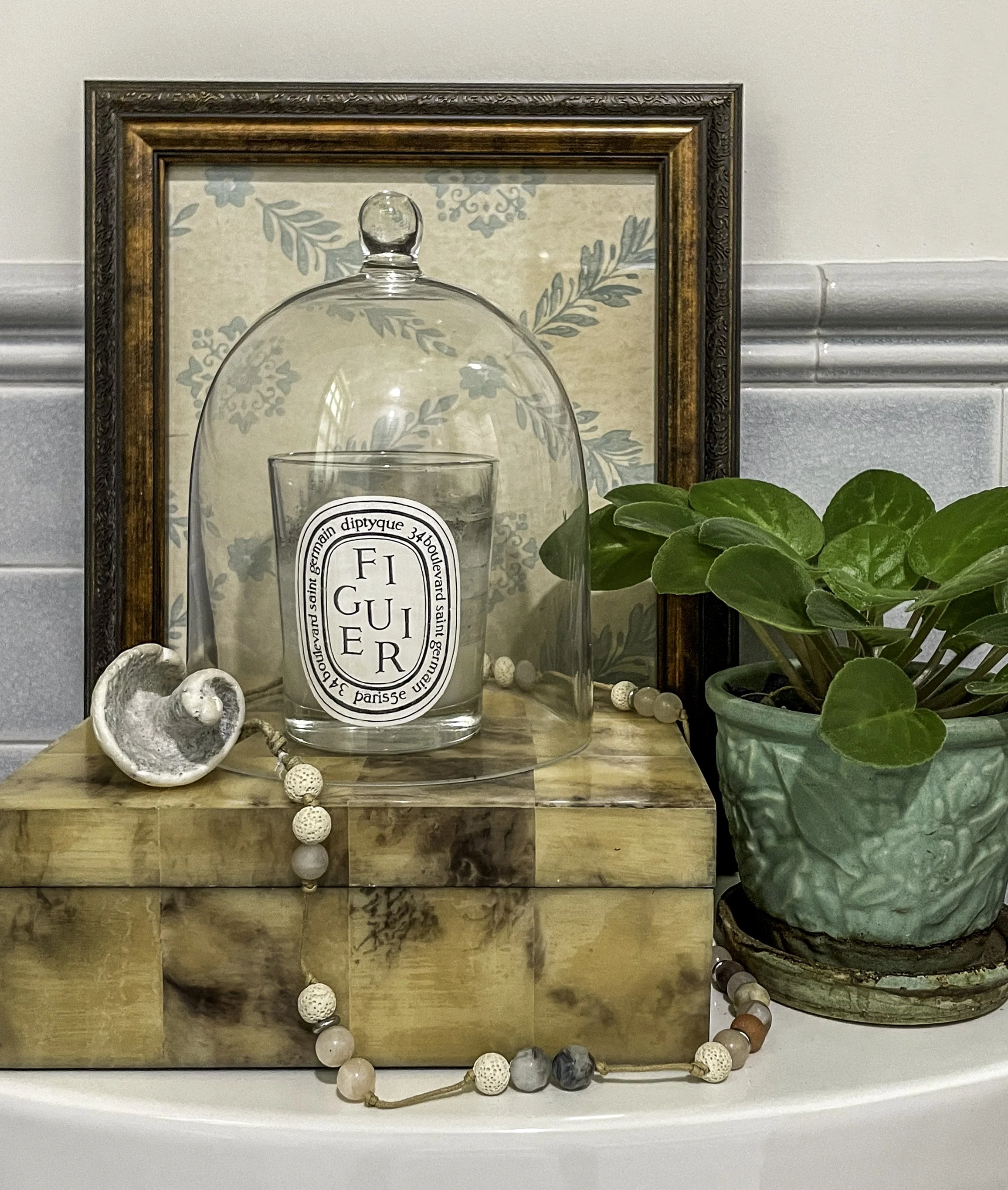 Styled bath vignette with a candle under a cloche, greenery, and framed botanical art