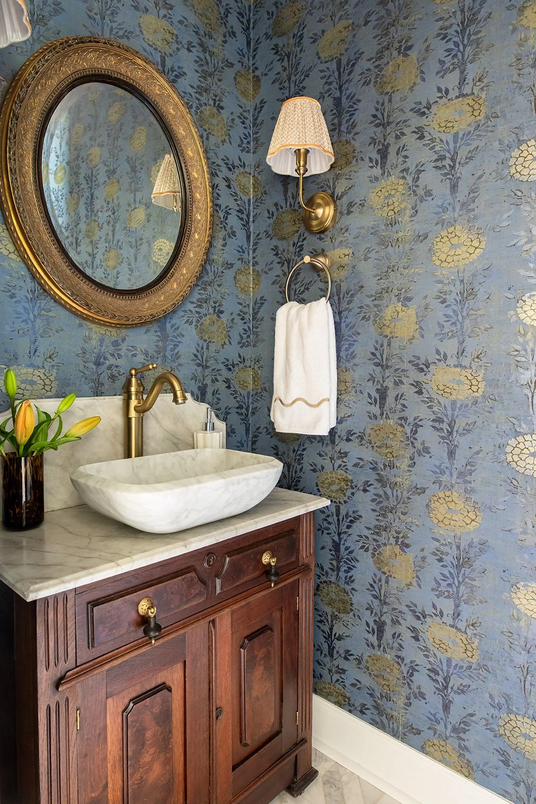 A vintage bathroom vanity with a marble countertop, white vessel sink, and brass fixtures. A vase of yellow tulips is on the left, and a soap dispenser is on the right. Above, a large oval gold-framed mirror and a wall-mounted brass sconce with a fabric lampshade are on a blue floral wallpapered wall. A white towel hangs on a brass towel ring.
