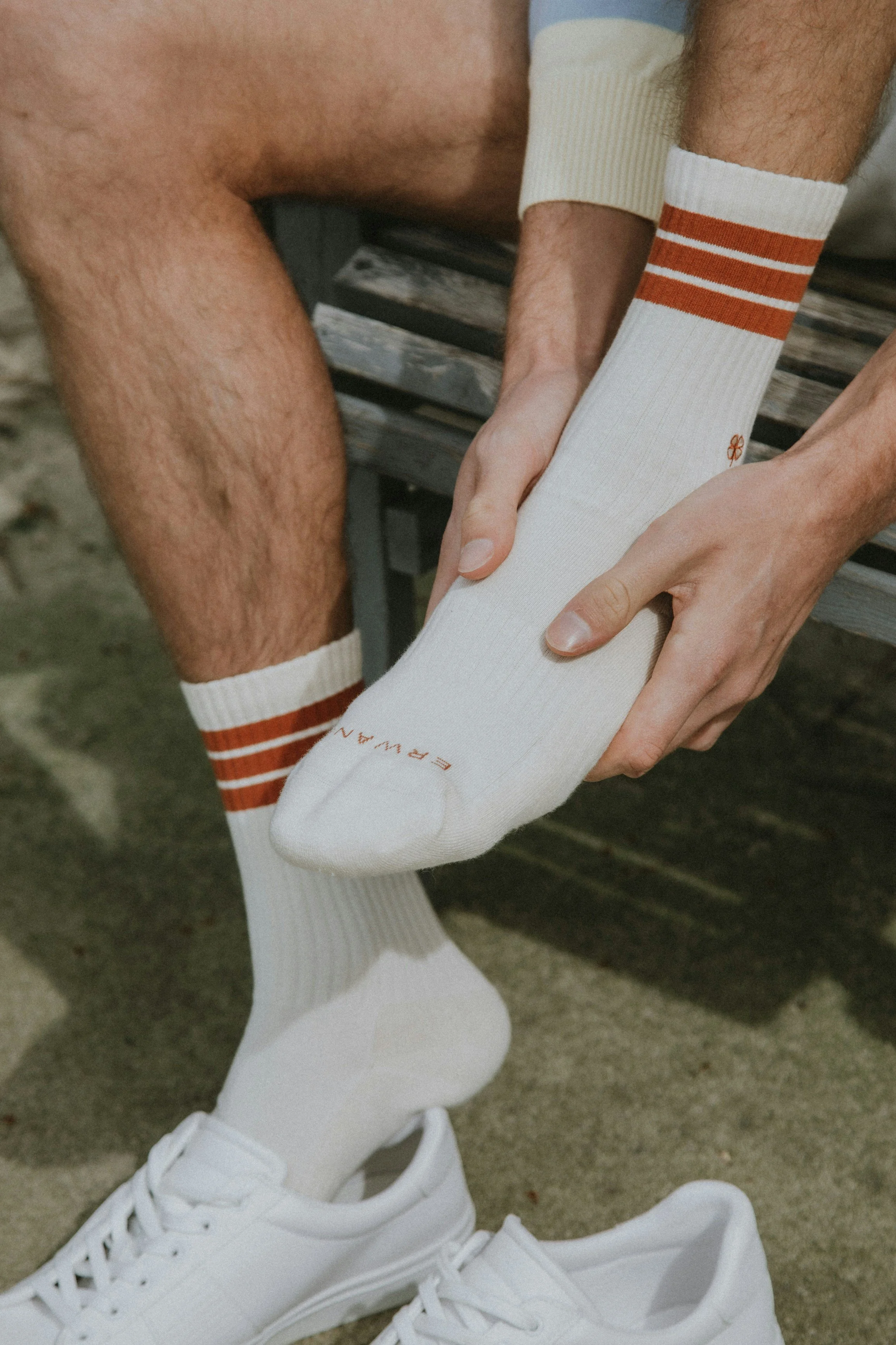 A person sitting on a bench wearing white socks with orange stripes and white sneakers, holding and adjusting their sock.