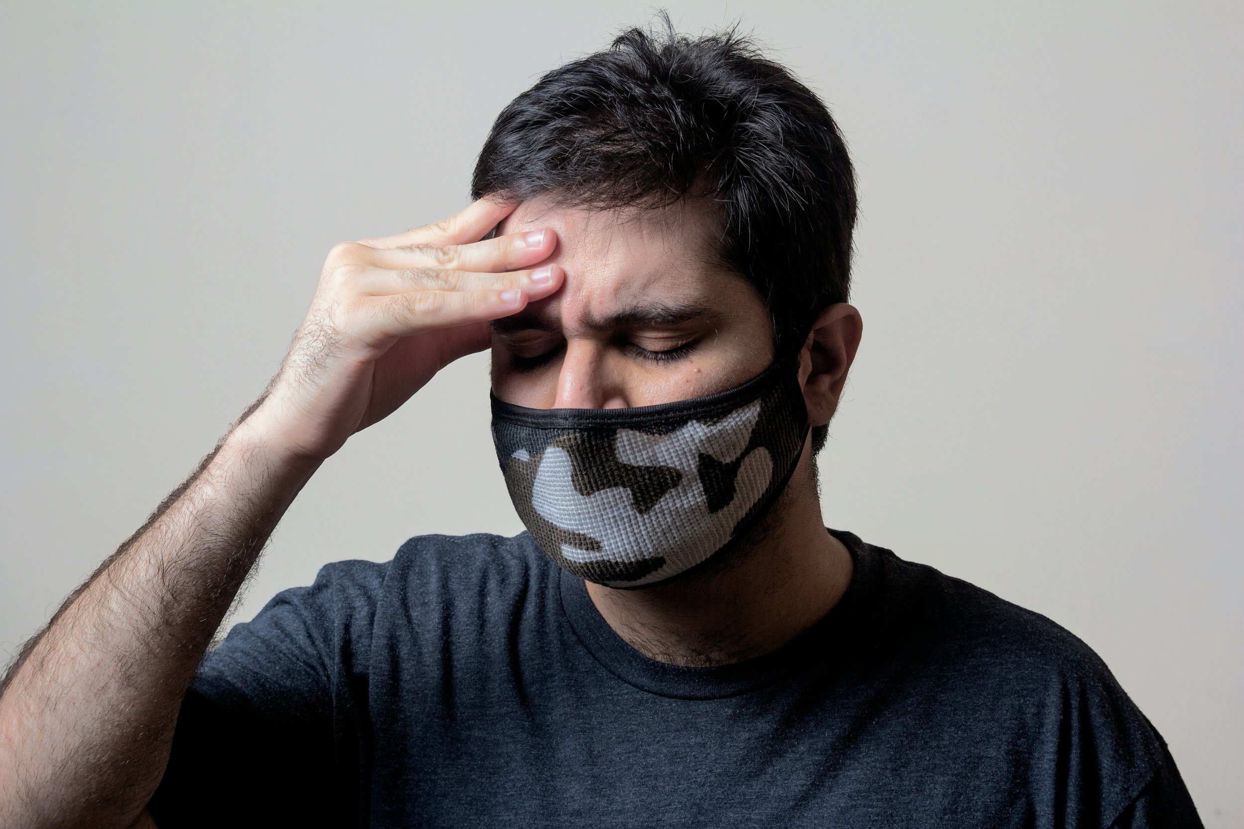 A man with dark hair, wearing a gray camouflage face mask and a dark gray t-shirt, holding his forehead with his right hand, appearing stressed or in pain.