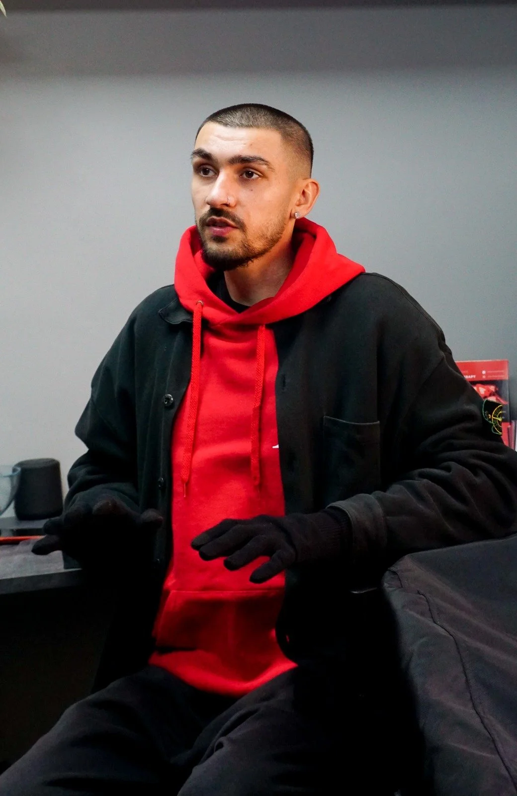 A man with short dark hair, a beard, and an earring wearing a red hoodie, black jacket, and black gloves, standing indoors against a plain gray wall.