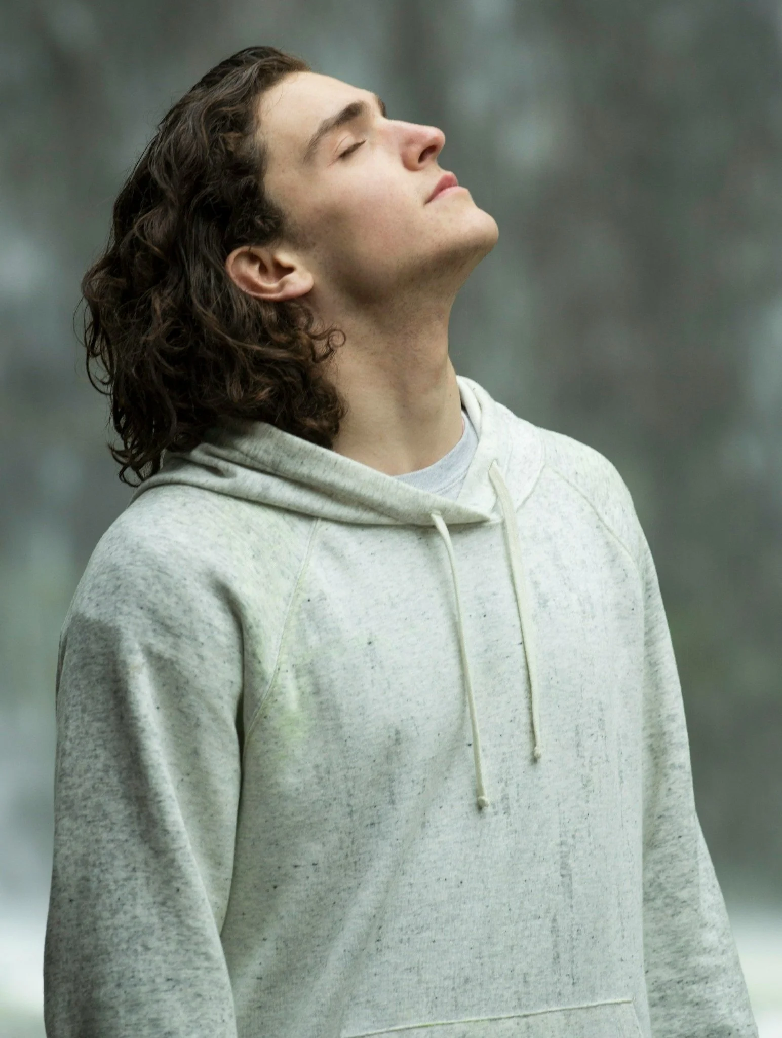 A young man with long, curly brown hair, wearing a light gray hoodie, stands outdoors with his eyes closed and face tilted slightly upward.