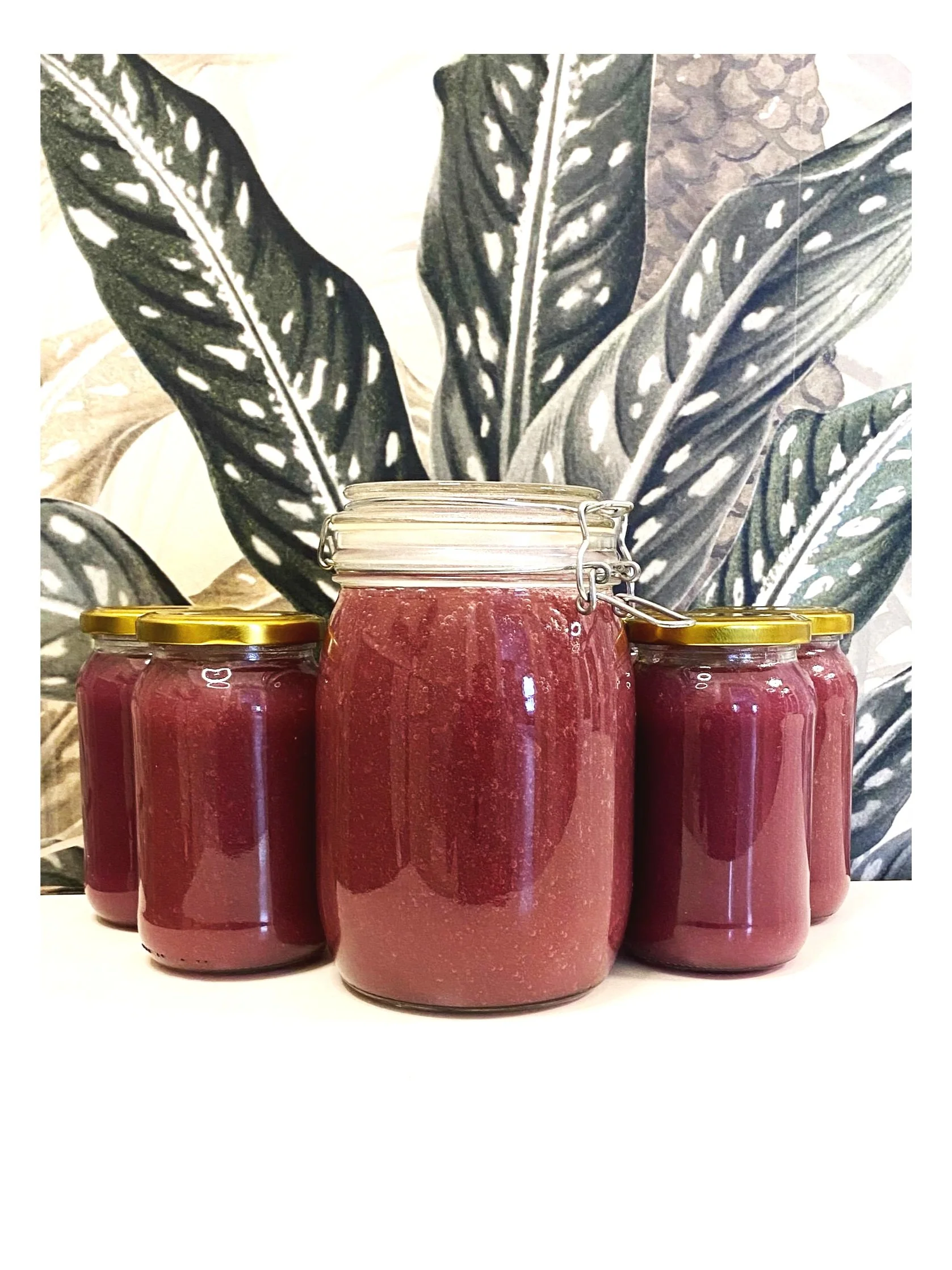 Four jars of homemade jam with gold lids, one larger jar in the center and three smaller jars surrounding it, in front of a black and white leafy plant wallpaper.